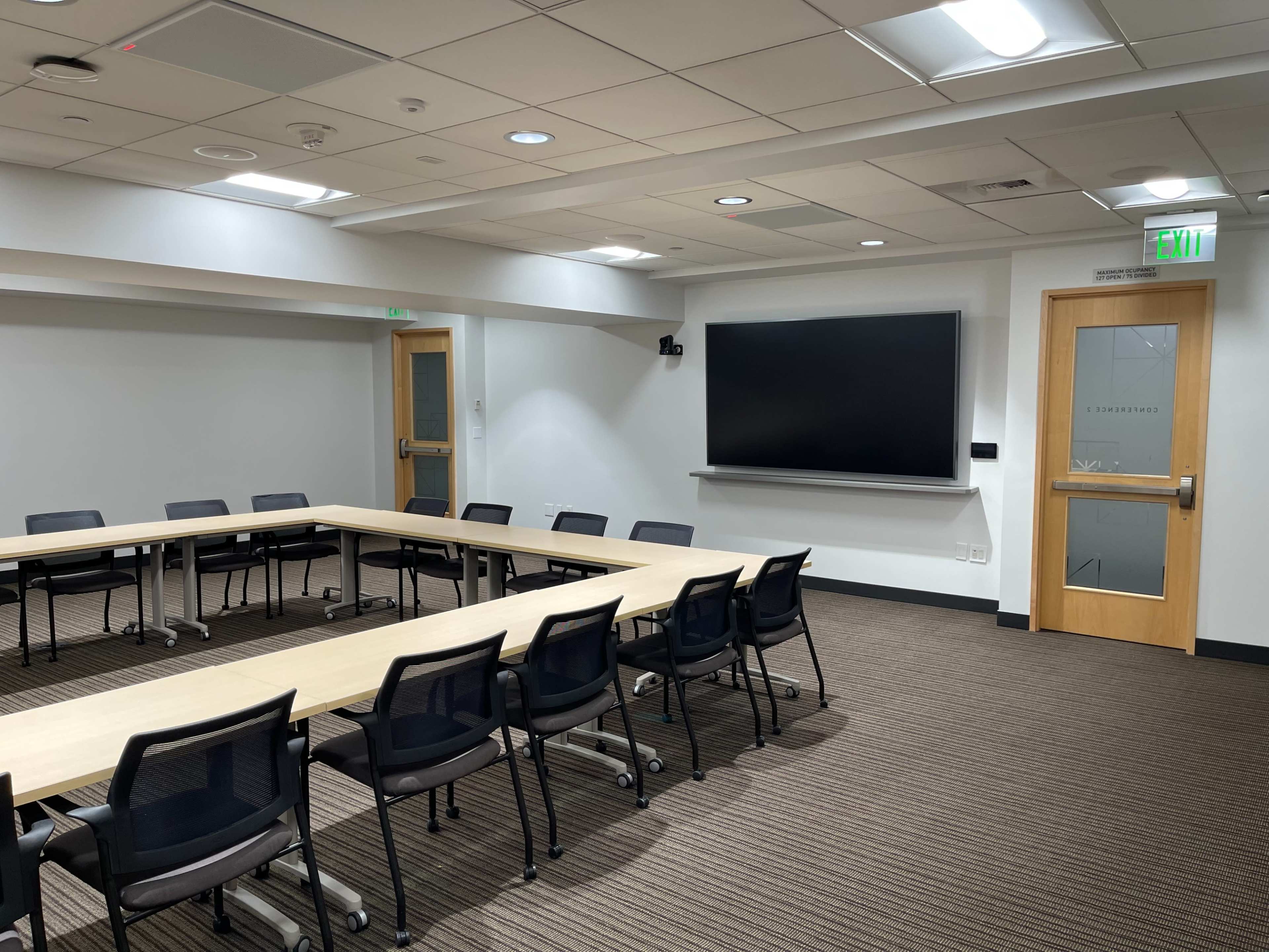 A conference room features a large rectangular table surrounded by chairs, with a wall-mounted screen and a door at one end.