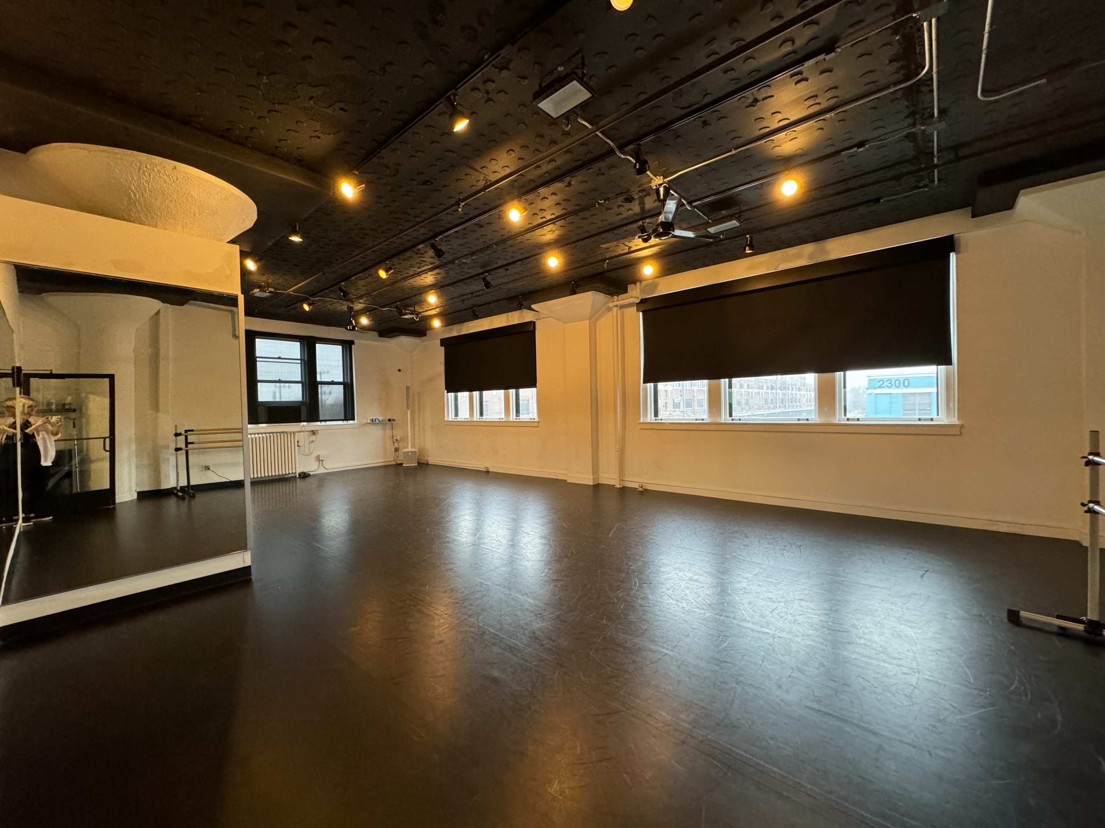 A spacious, well-lit dance studio with hardwood flooring, large windows covered by black blinds, and a mirrored wall.