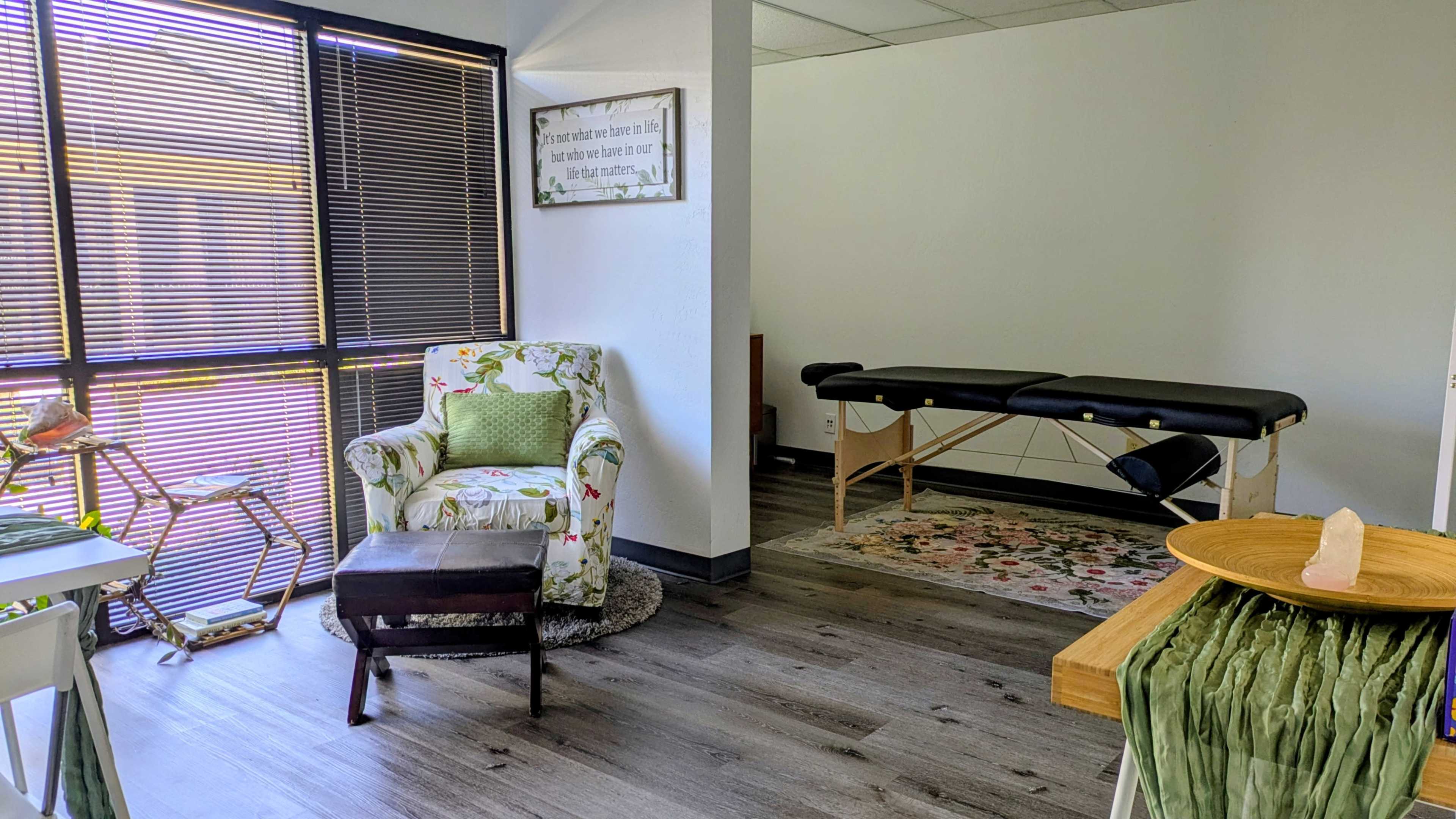 The image shows a therapy room featuring a floral chair, a massage table, and a decorative rug on a wooden floor.