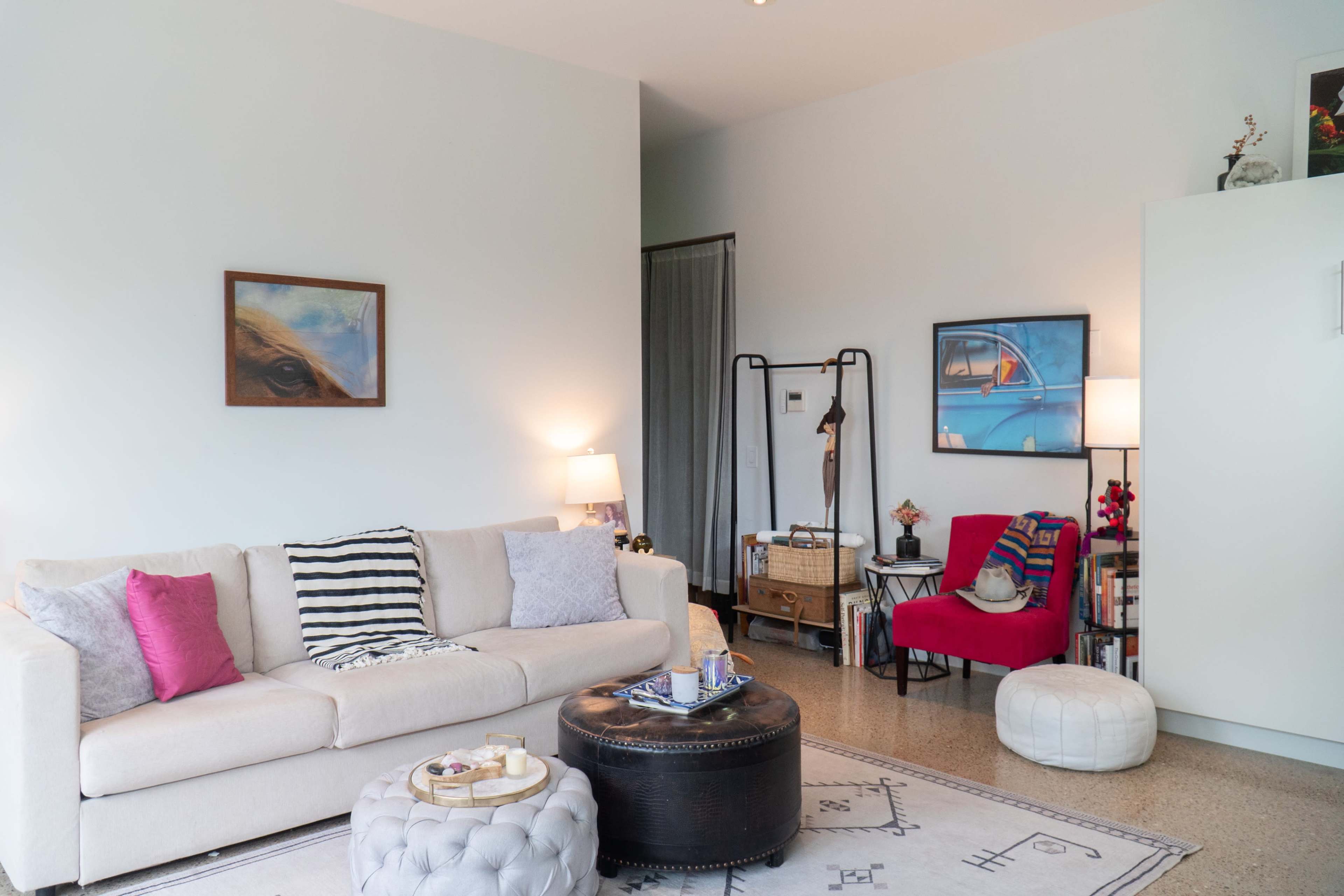 The living room features a beige sofa with colorful cushions, a black coffee table, a light-colored pouf, and artwork on the walls, with a mix of lighting from lamps.