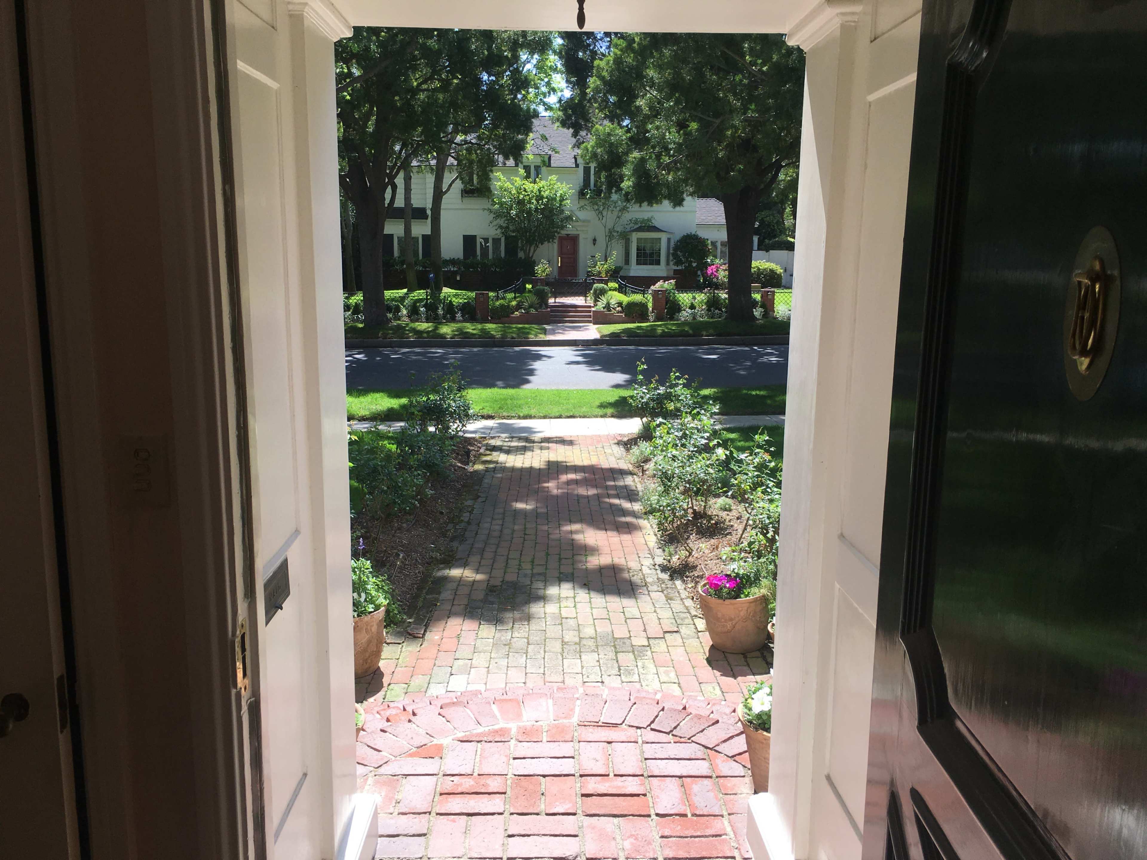 The view from an open front door shows a brick pathway leading to a house across the street, surrounded by trees and landscaped gardens.
