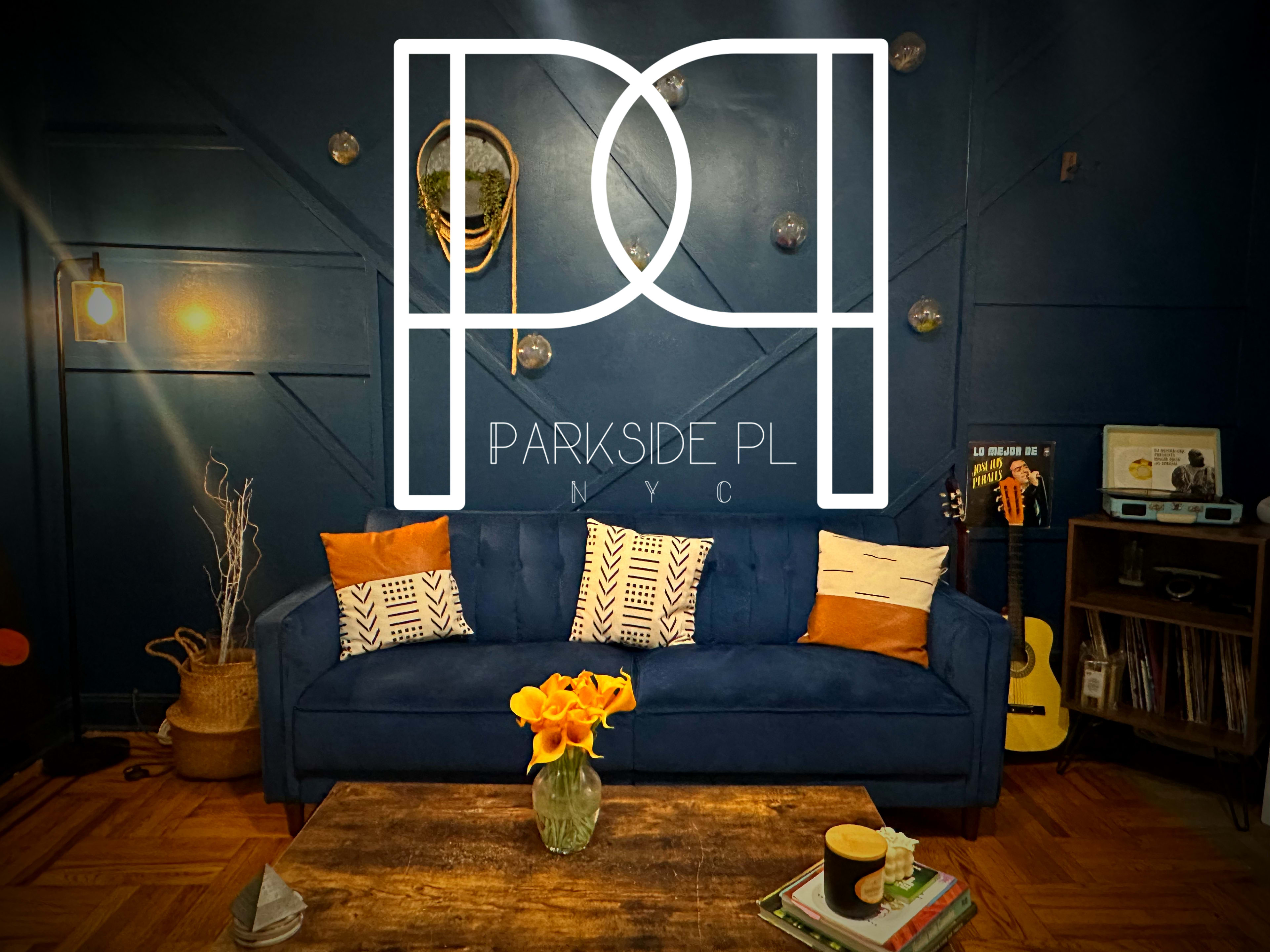 The image shows a cozy living room with a blue couch adorned with orange and patterned cushions, a wooden coffee table with a vase of yellow flowers, and a decorative wall featuring a mix of textures and lighting.