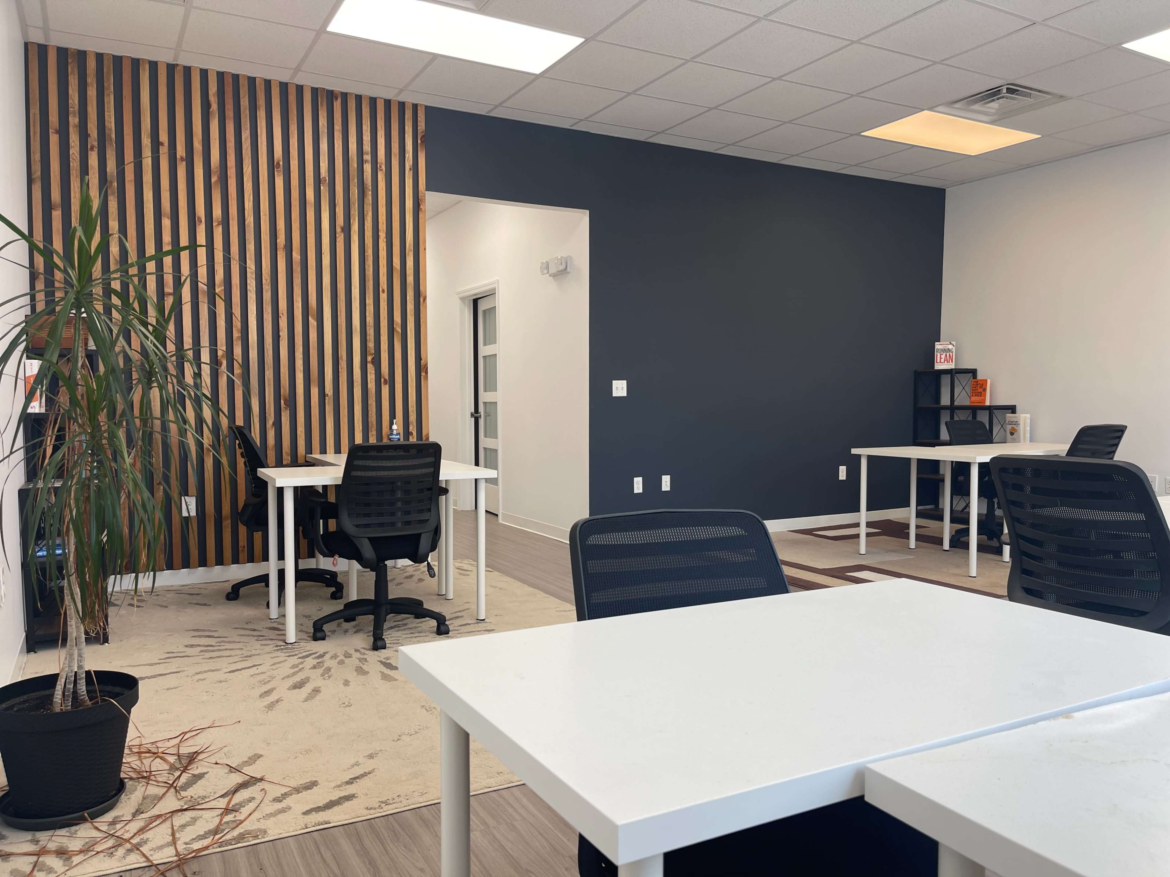 The image shows a spacious office with white tables and black chairs, featuring a wooden accent wall and a potted plant in the corner.