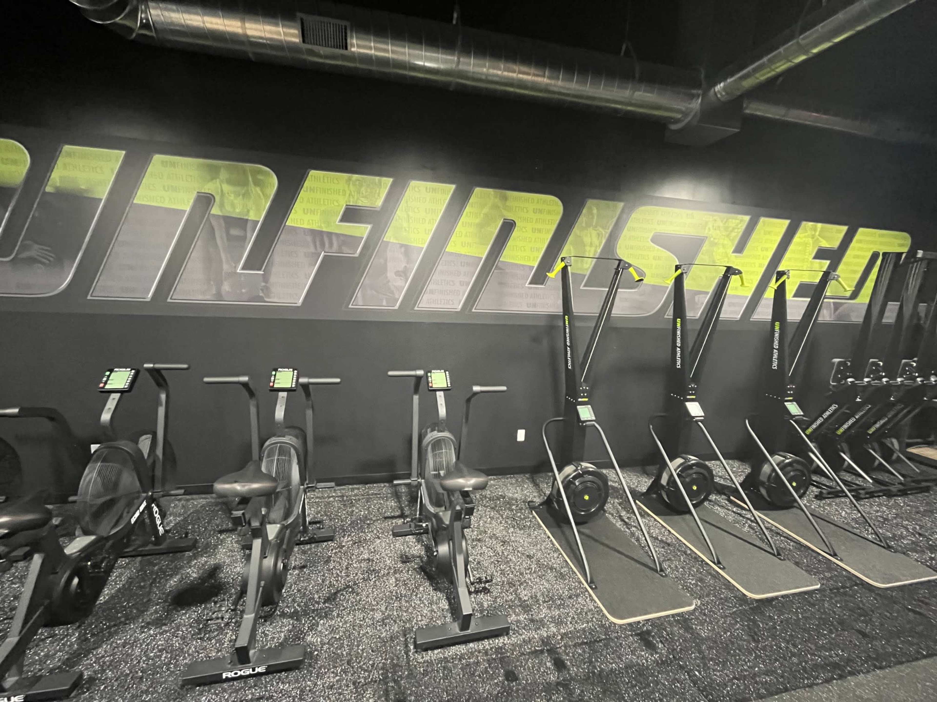 The image shows a gym interior featuring rowing machines and exercise equipment lined up against a dark wall with the word "UNFINISHED" displayed in bold letters.