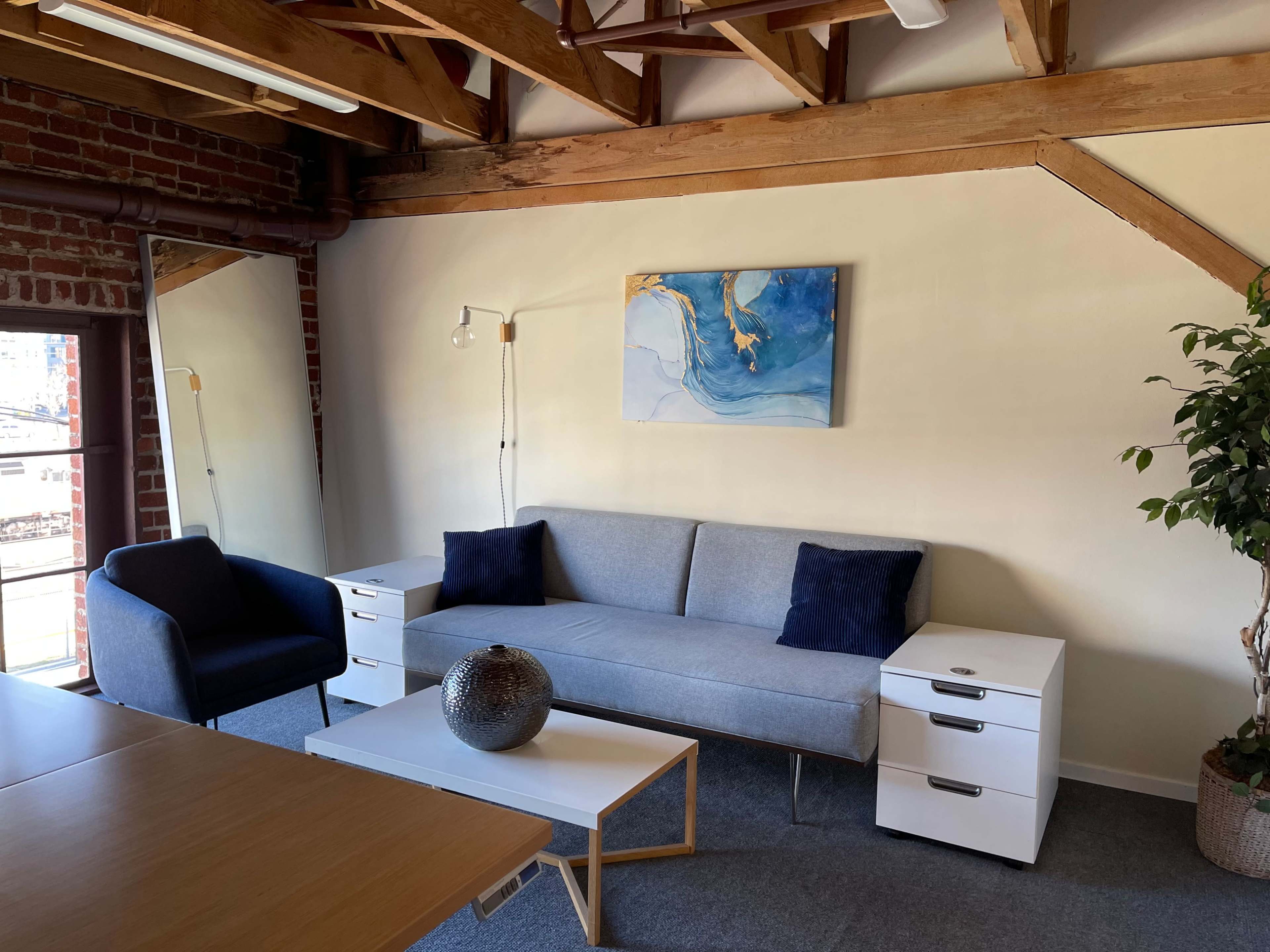 A modern living space with a gray sofa, two white side tables, a blue accent chair, and a large wall-mounted painting, set against a backdrop of exposed wooden beams and brick walls.