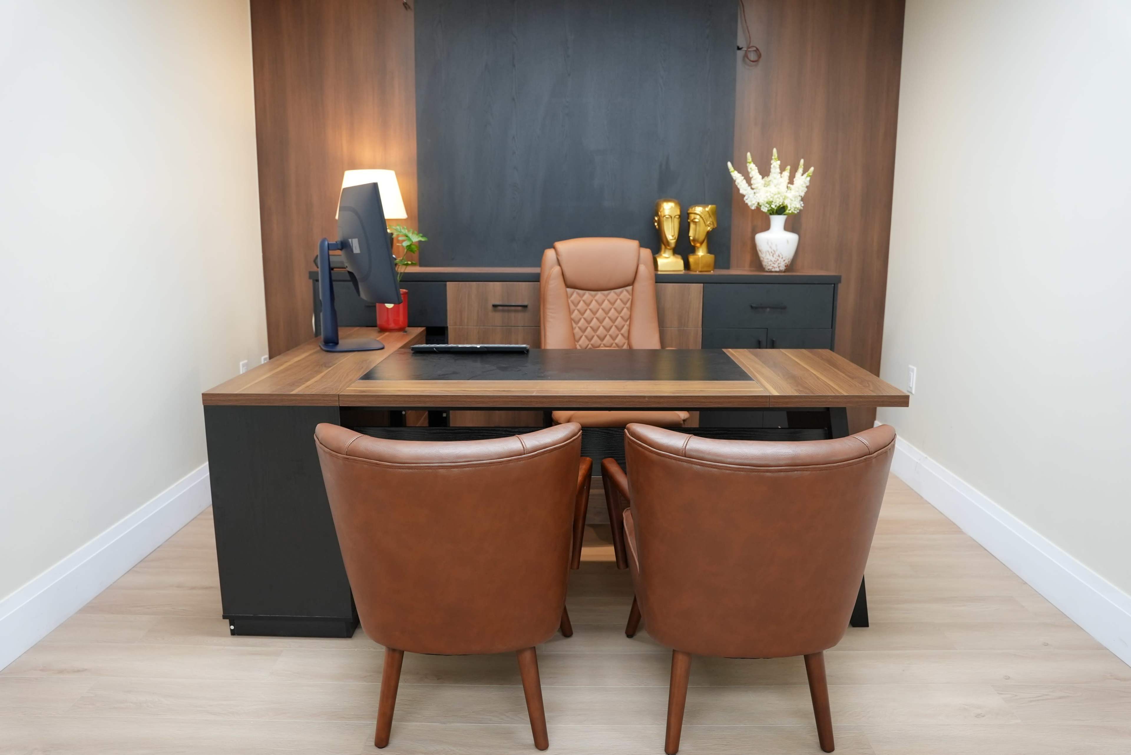 A modern office features a wooden desk with a computer monitor, two brown leather chairs, and decorative items on a dark panel backdrop.