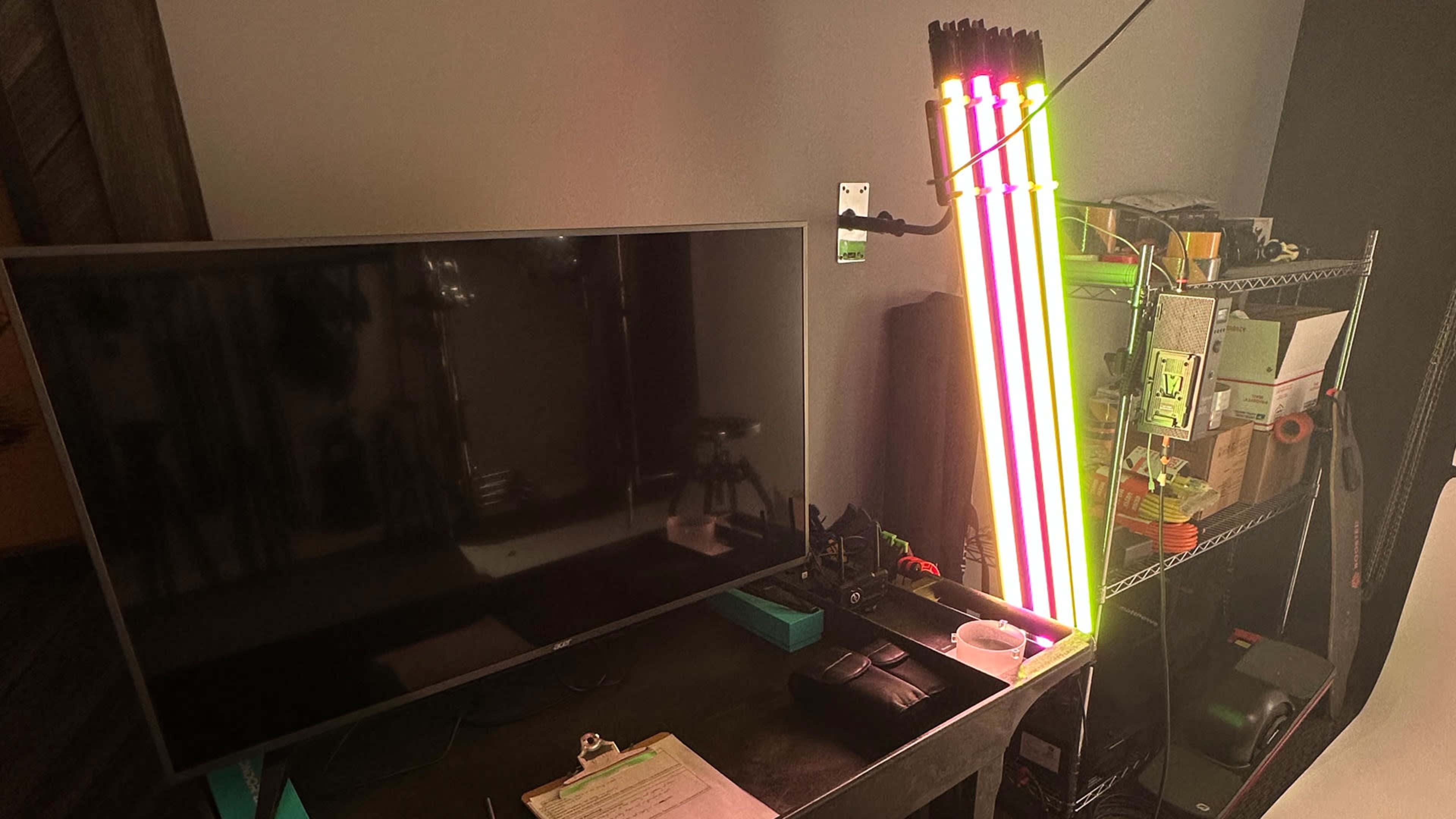 A television is positioned on a dark desk next to a set of colorful LED light tubes and various items on shelves in the background.
