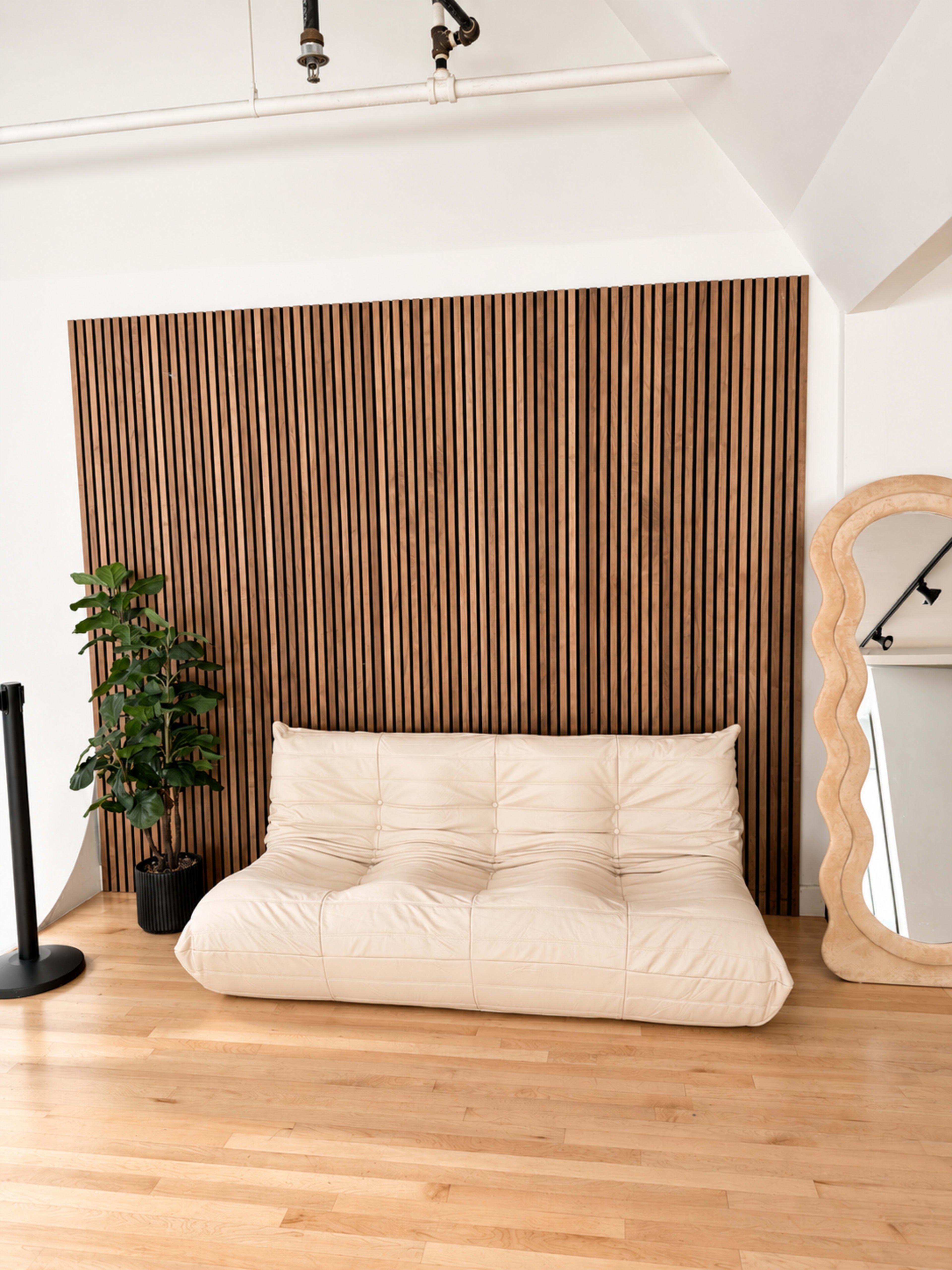The image features a light-colored sofa against a wooden slatted wall, accompanied by a potted plant and a decorative mirror.
