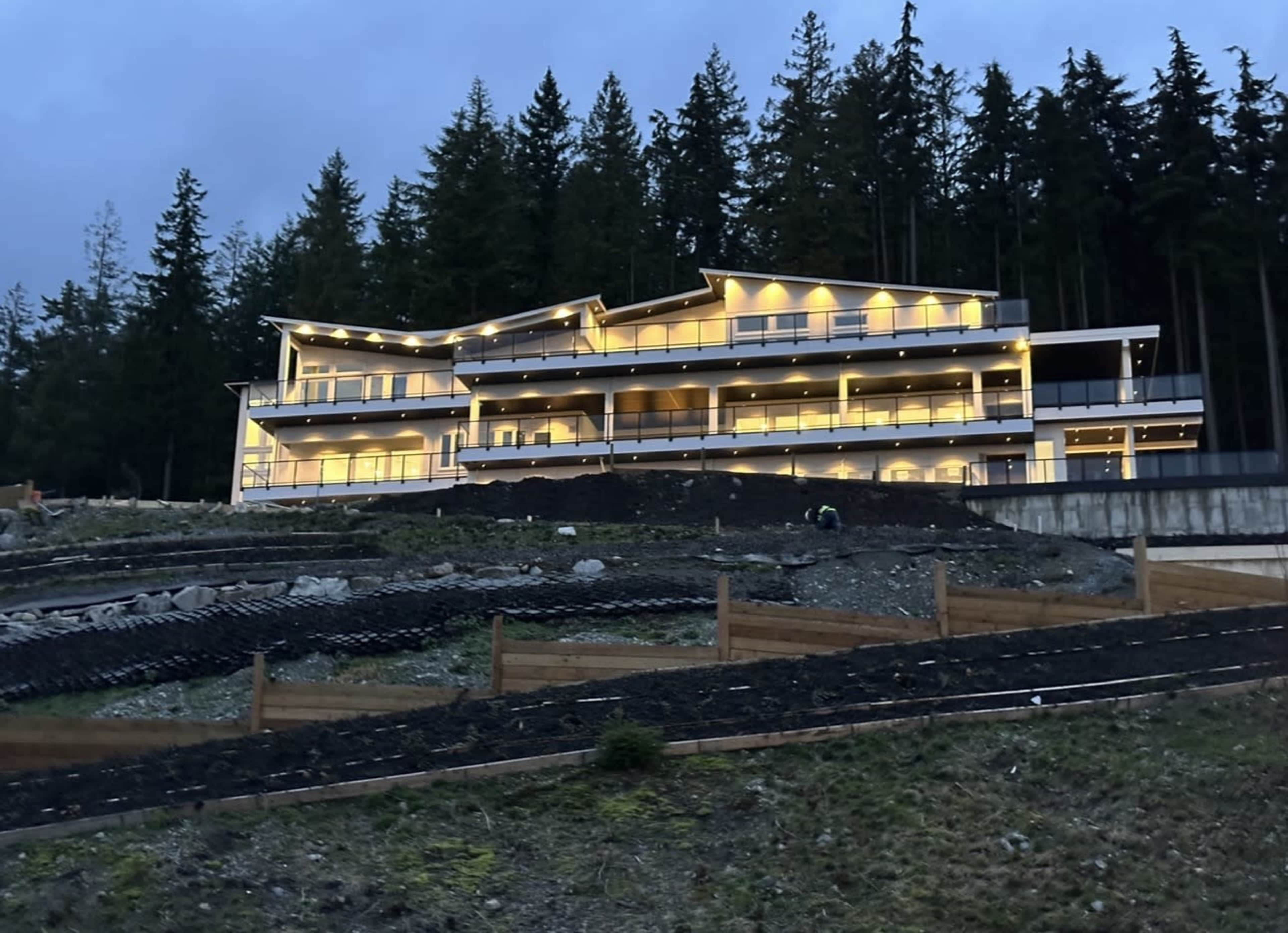 A modern multi-story building illuminated at dusk is set against a backdrop of tall trees.