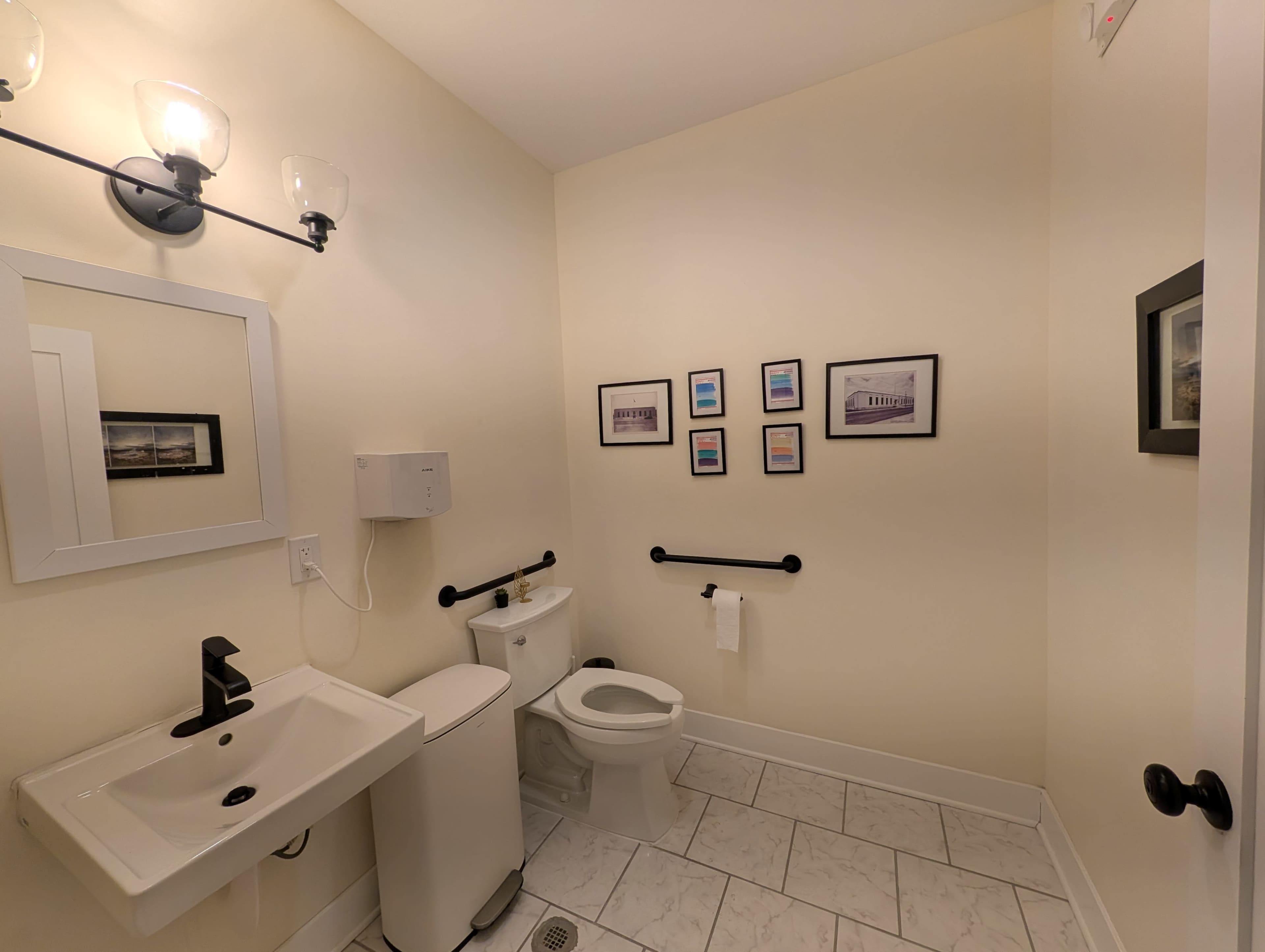 The image shows a small bathroom featuring a white toilet, a sink with a modern faucet, and several framed pictures on the wall.