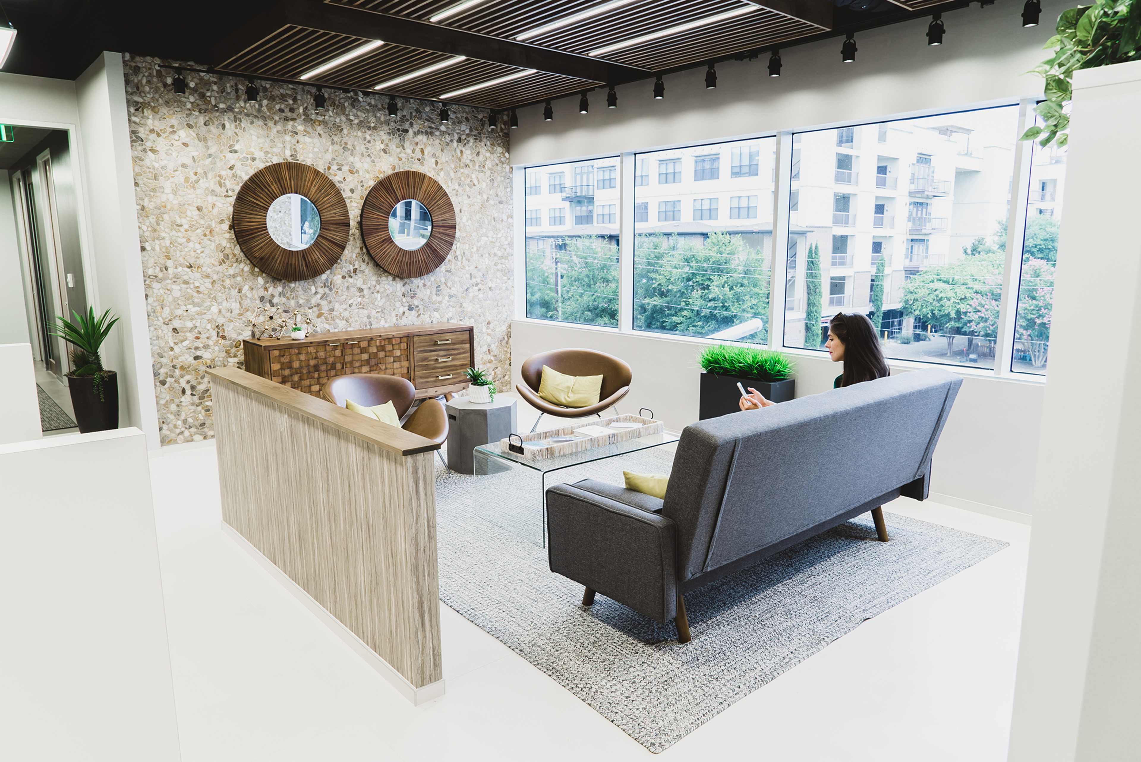 A modern reception area with a gray sofa, two brown chairs, decorative mirrors, and a large window overlooking an outdoor view.