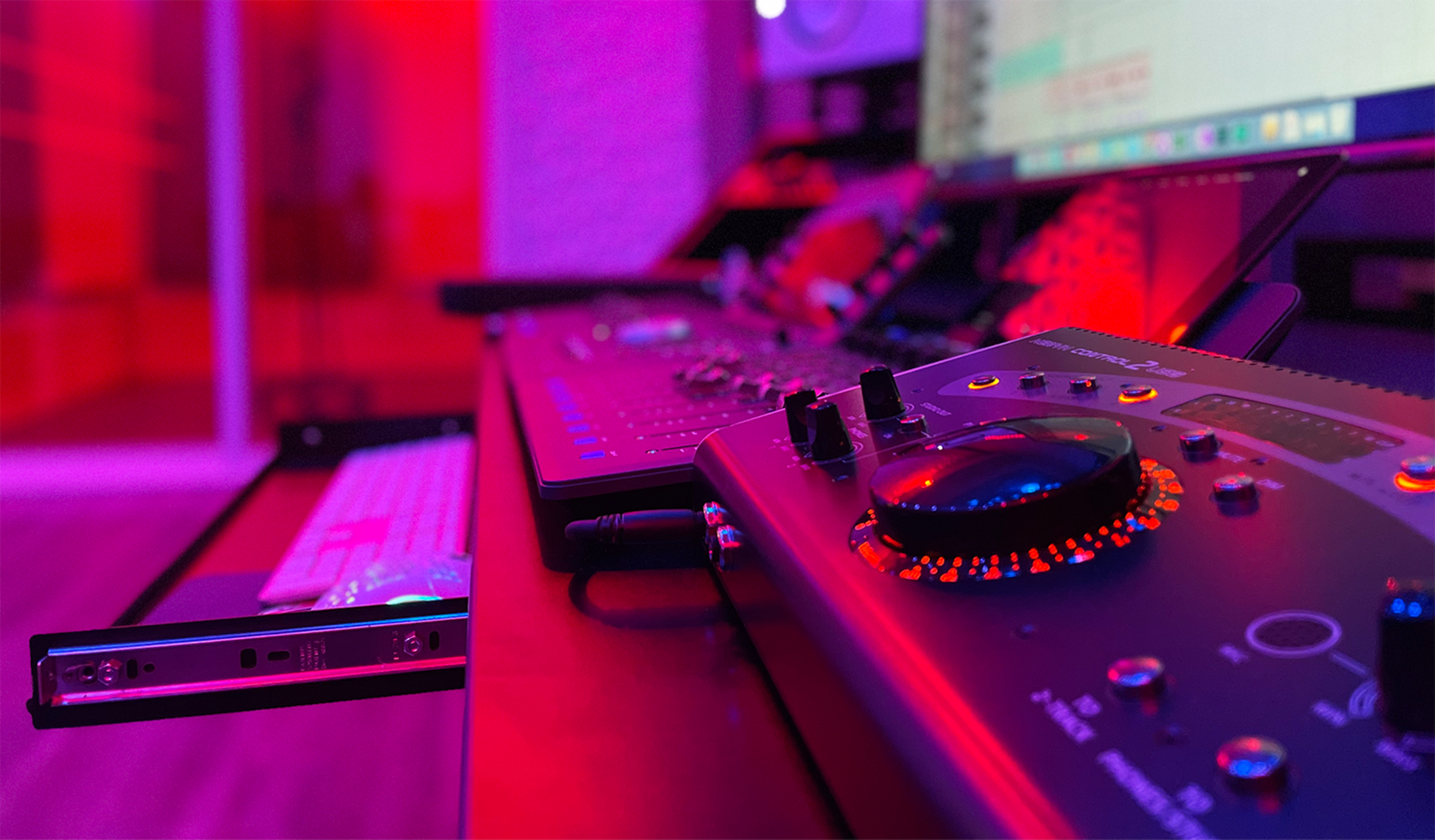 A close-up view of a recording studio workspace featuring a digital audio interface, a keyboard, and a computer screen illuminated by red lighting.