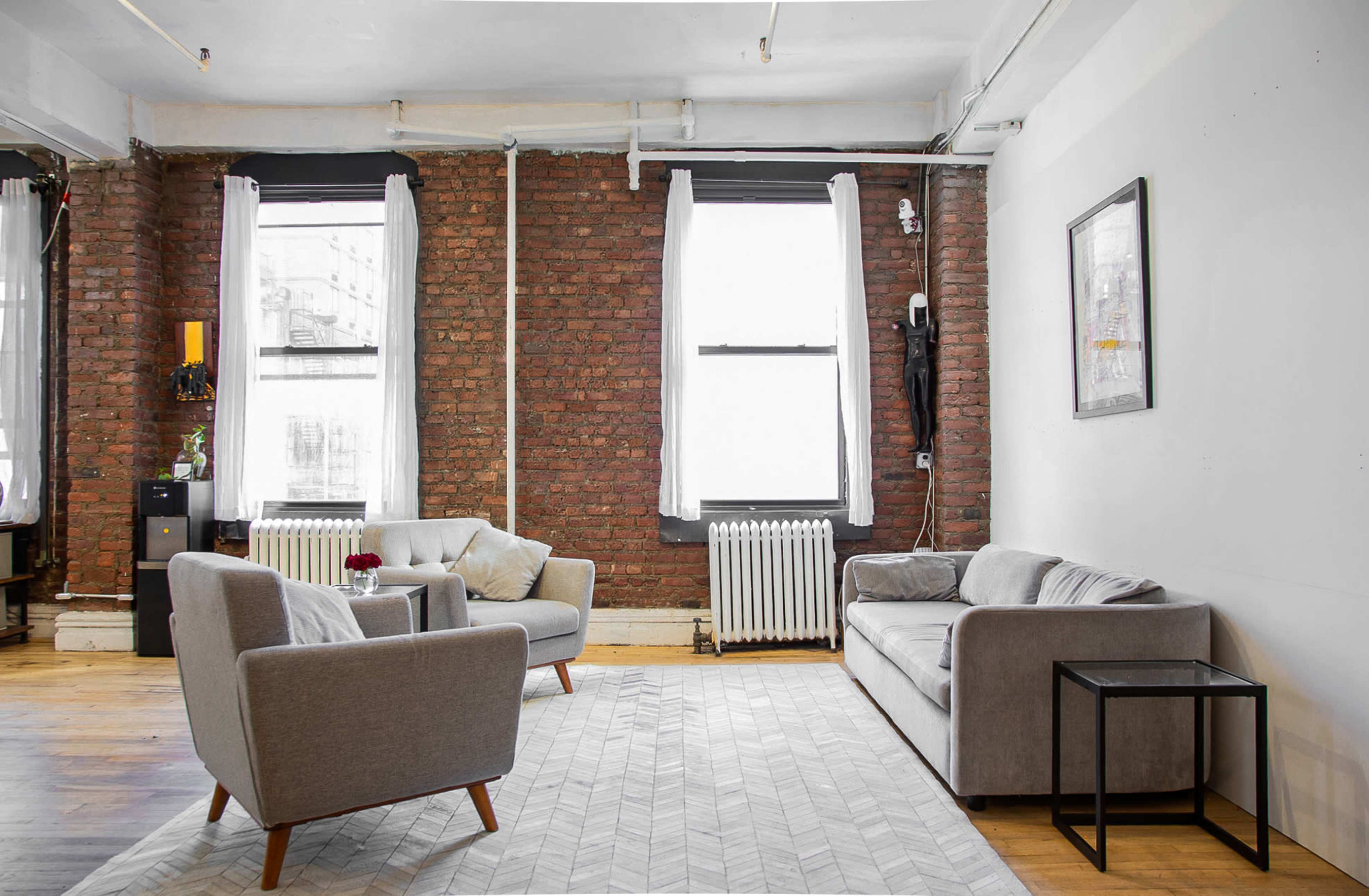 A modern living room features exposed brick walls, two gray armchairs, a gray sofa, a small black side table, and large windows with white curtains.