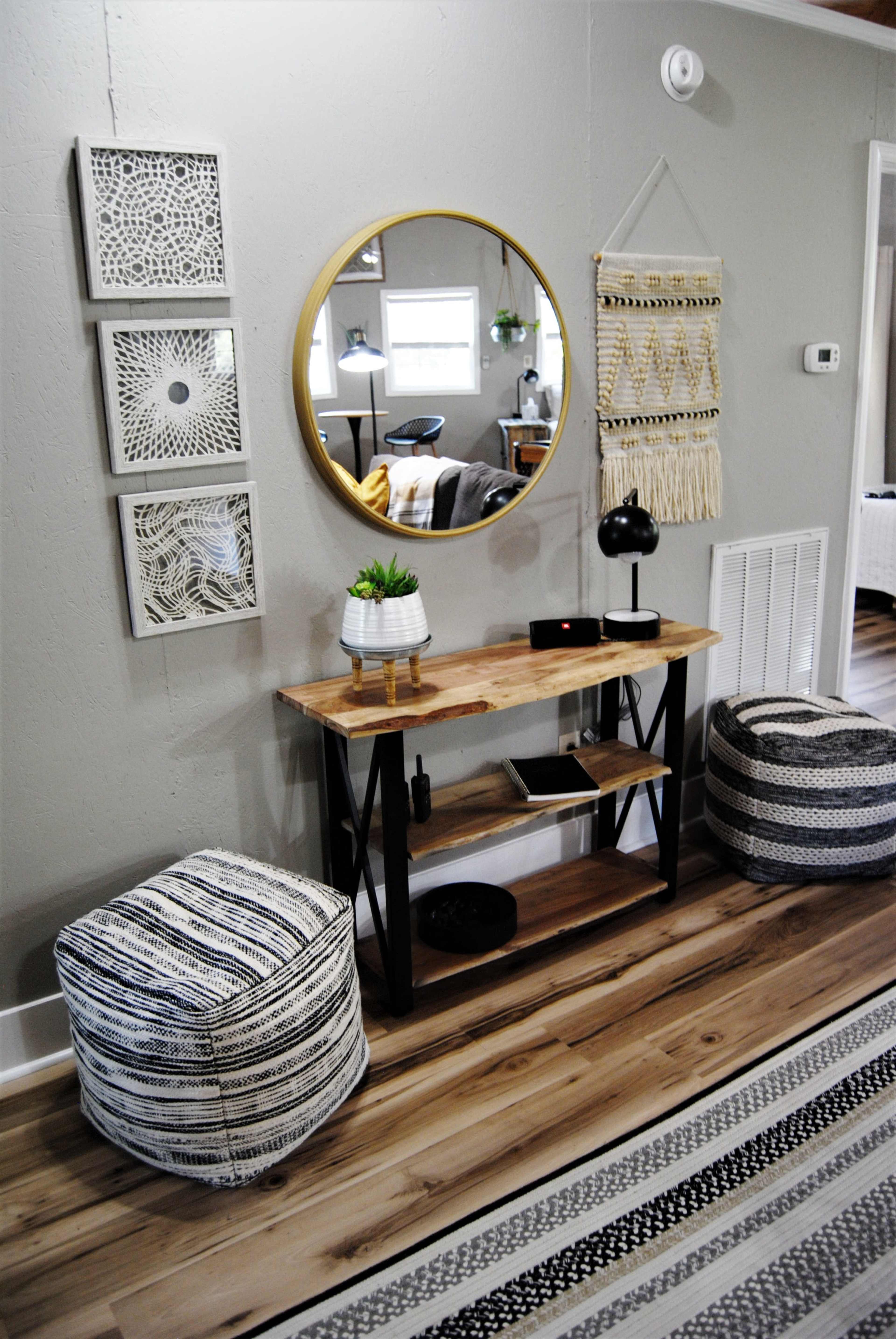 A modern entryway with a round mirror above a wooden console table, flanked by decorative wall art and accompanied by two patterned poufs.