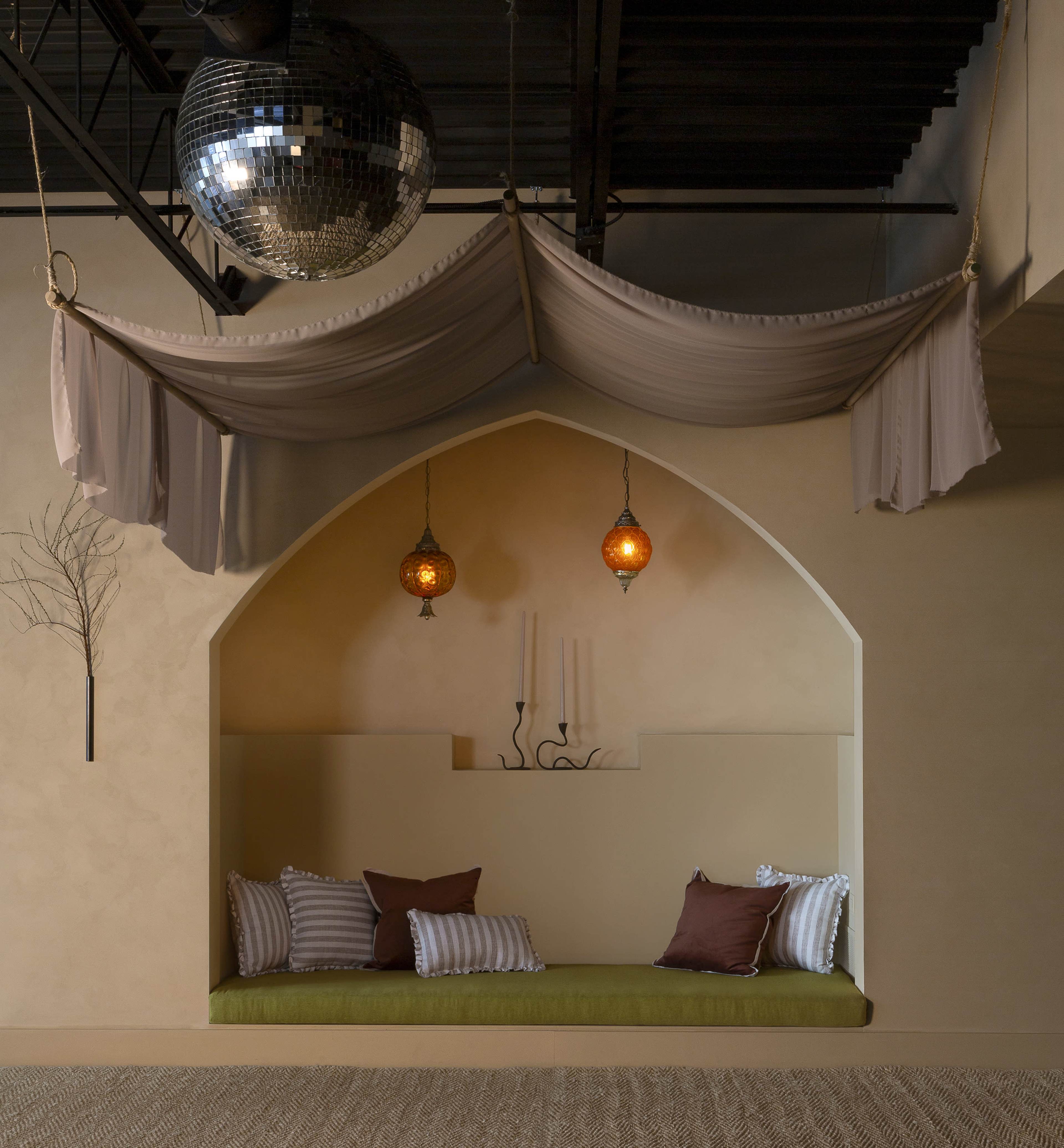 The image features a cozy alcove with a green seating area adorned with pillows, flanked by geometric lanterns and draped fabric, set against a soft, neutral wall.