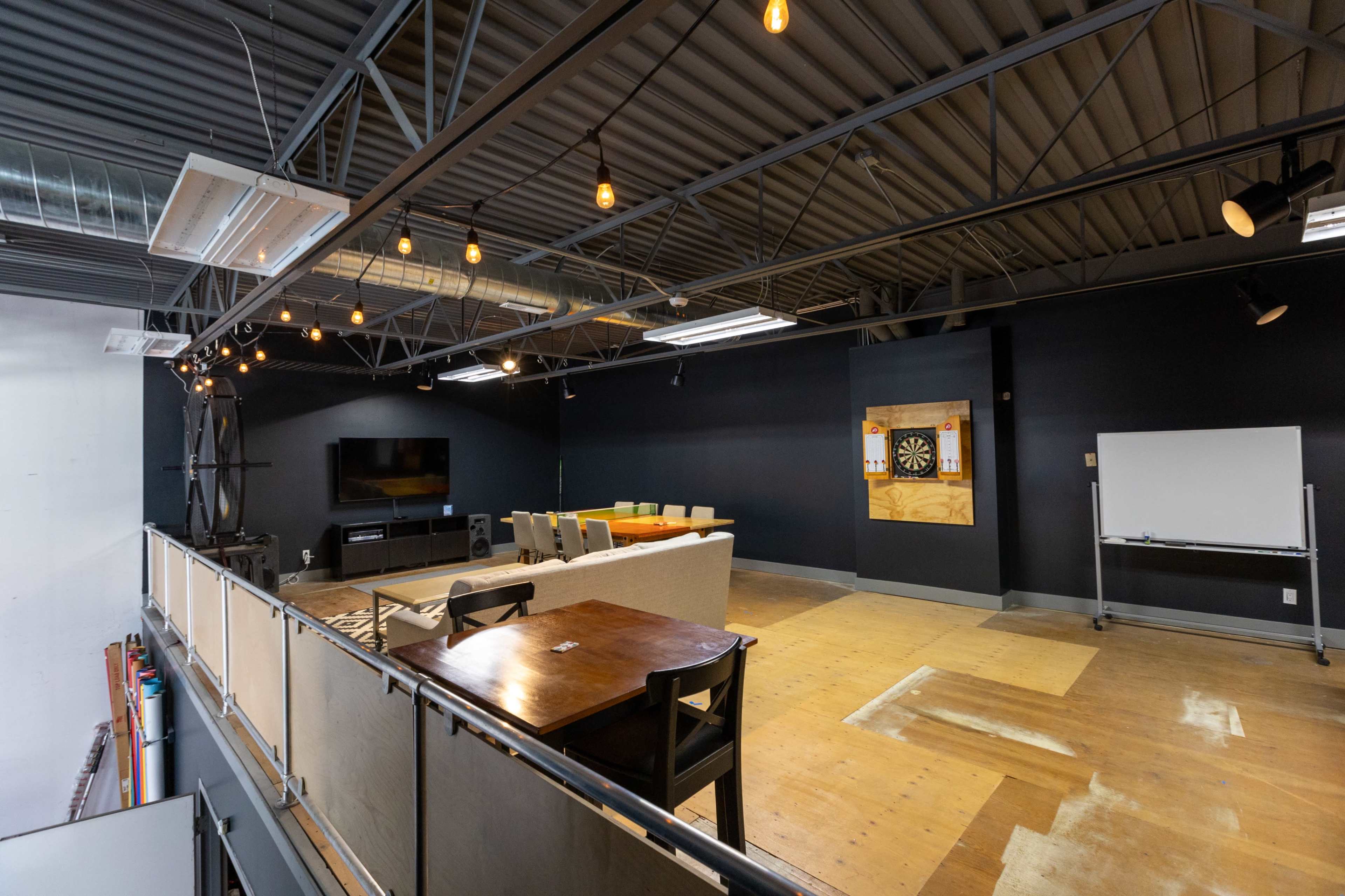 An interior space with a second-floor balcony overlooking a seating area, a dartboard, and a large television in a modern decor.