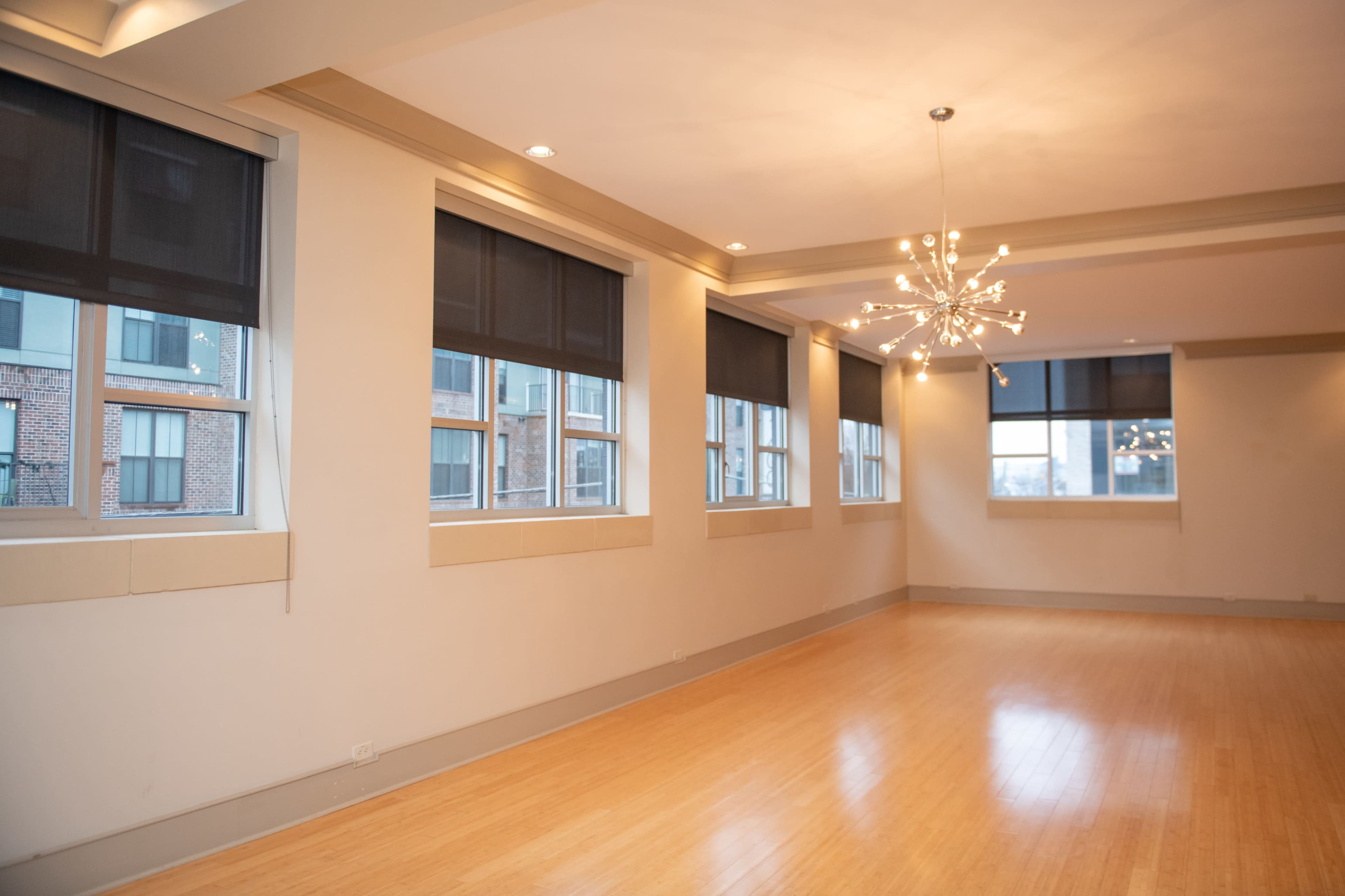 The image shows a spacious room with large windows and a modern chandelier hanging from the ceiling, featuring wooden flooring.