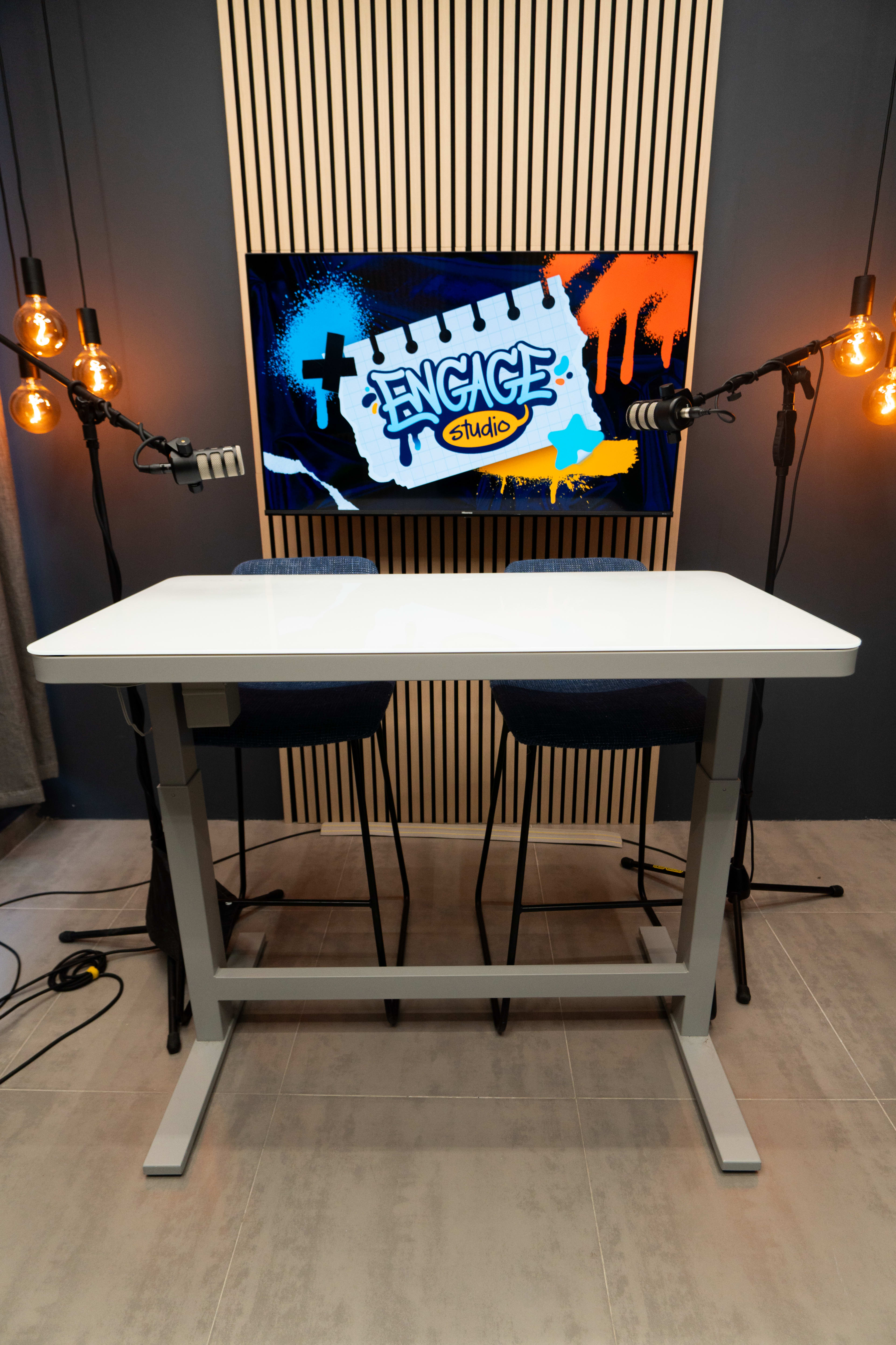The image shows a modern studio setup with a white table, two blue chairs, and a large screen displaying the "ENGAGE Studio" logo, surrounded by decorative lighting.