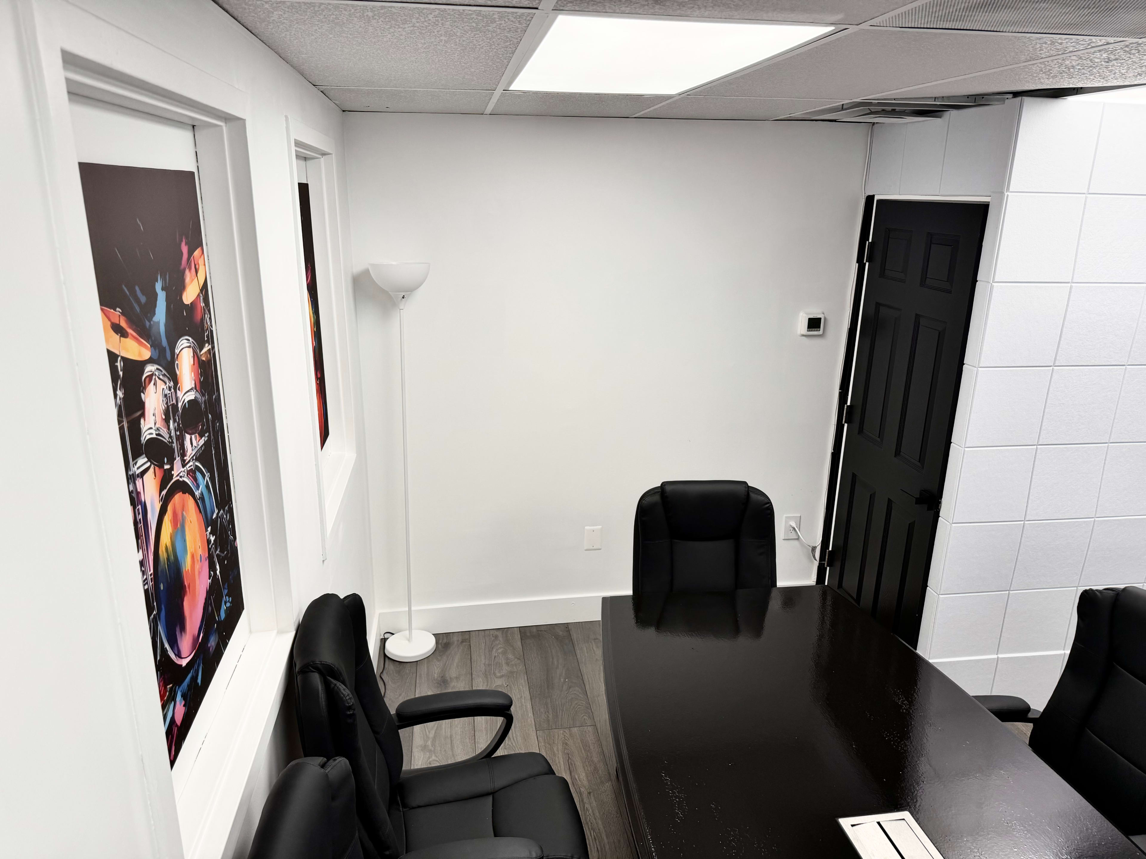 The image shows a conference room with a black table, several black chairs, a floor lamp, and artwork of a drum set on the walls.