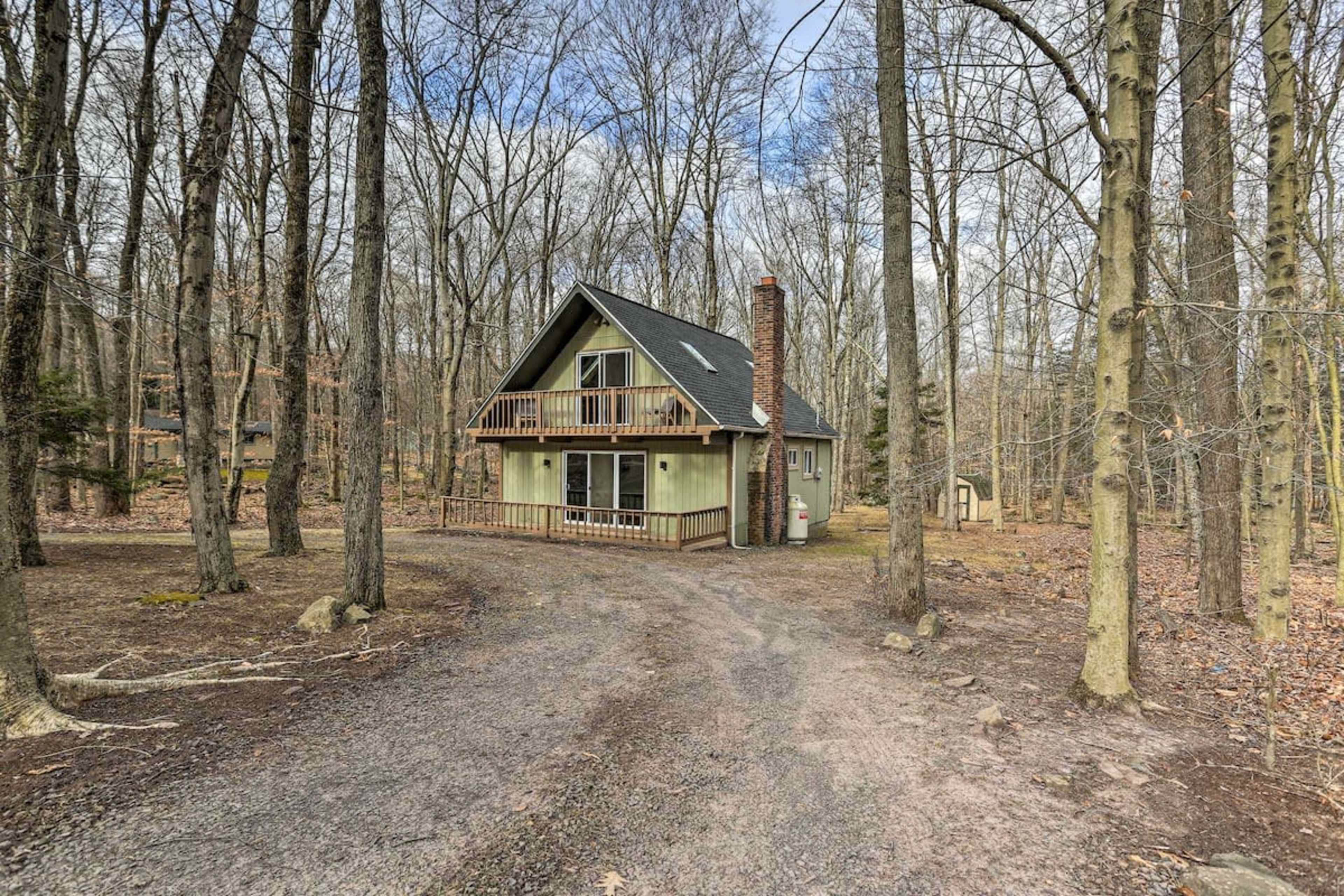 A two-story, green house with a sloped roof and a front porch is set among bare trees in a wooded area.