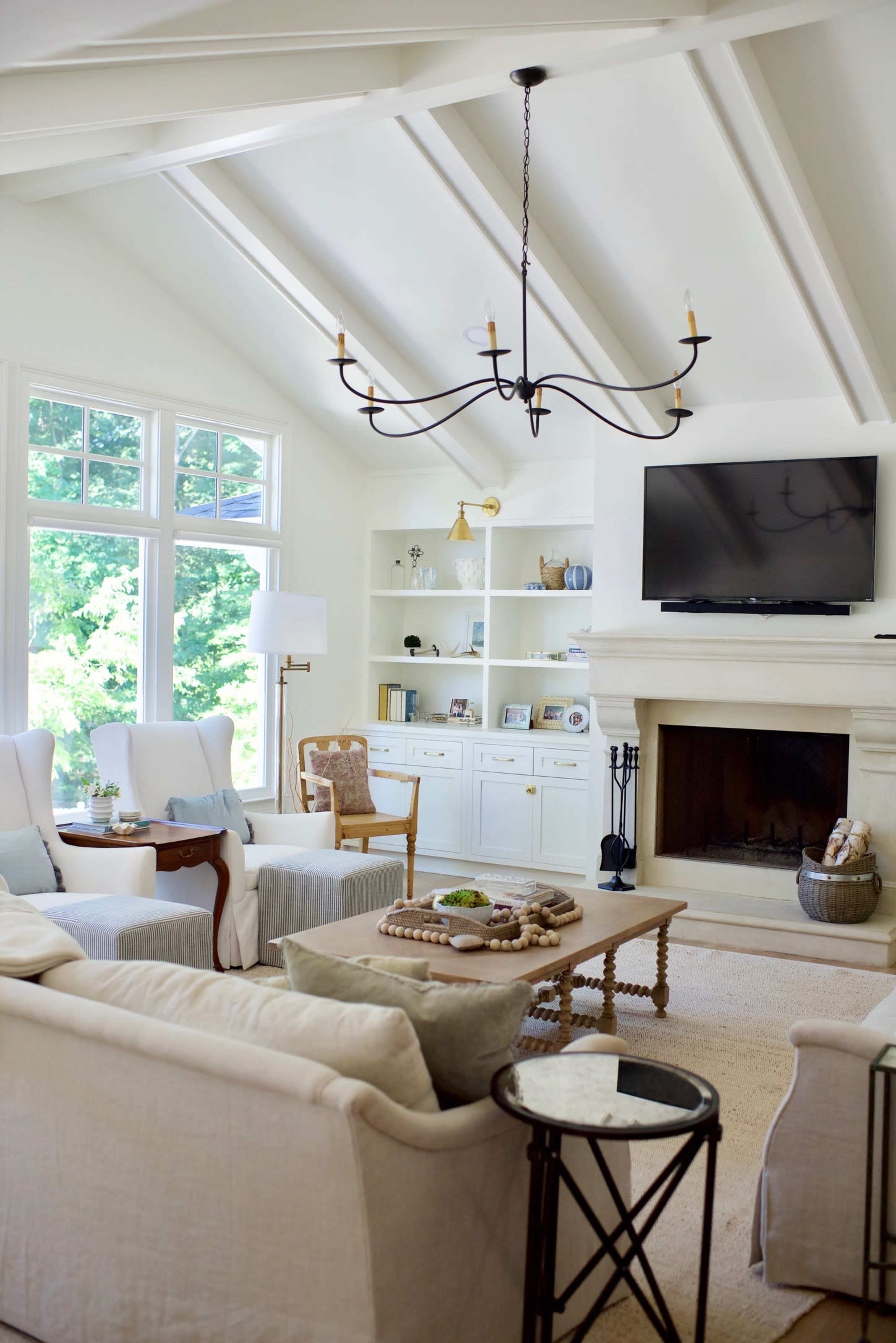 A bright living room features a white color scheme with a modern chandelier, a television mounted above a fireplace, and comfortable seating arranged around a coffee table.