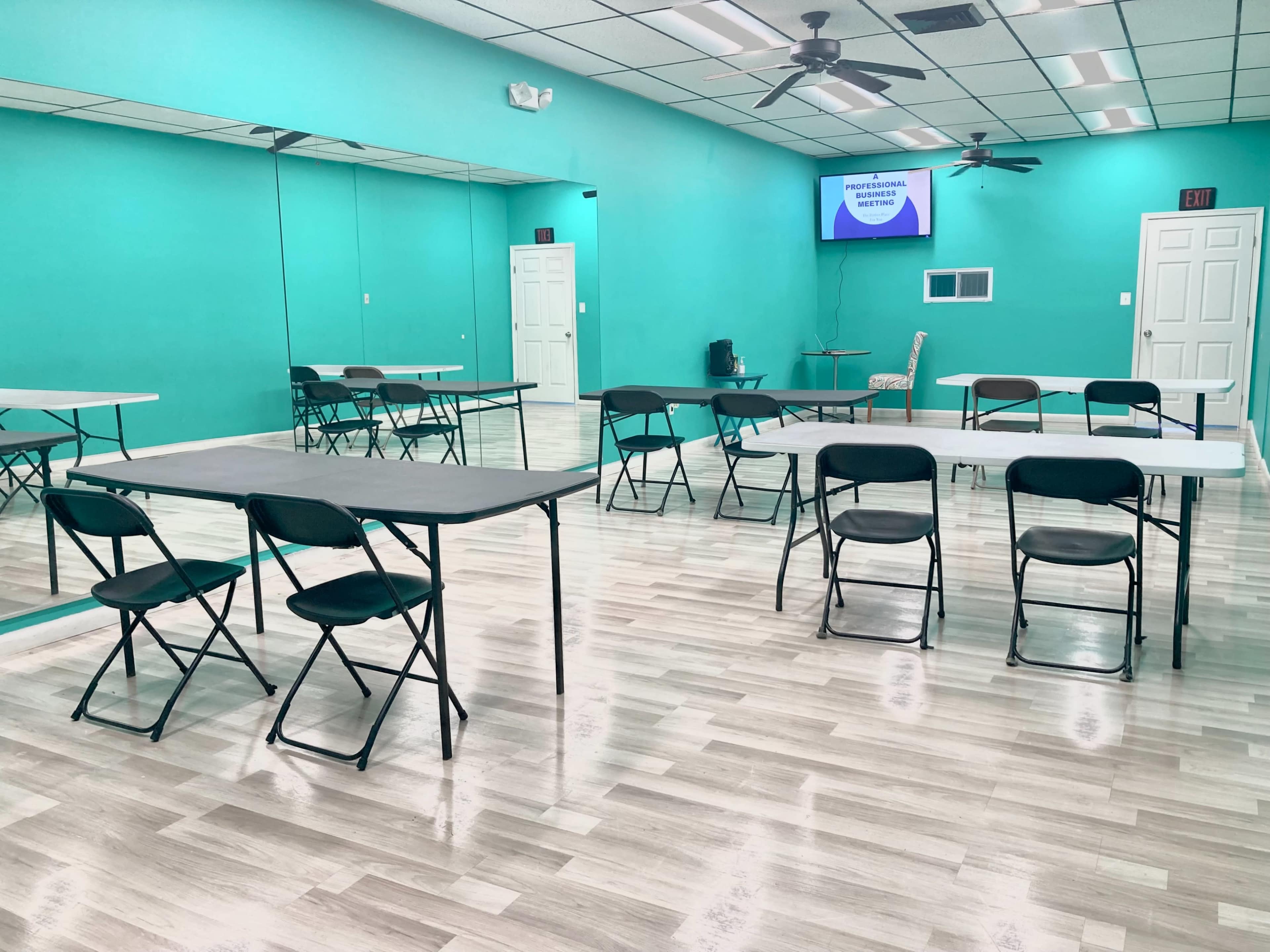 A brightly colored room with teal walls, featuring several folding tables and chairs arranged throughout, and a large mirror on one side.