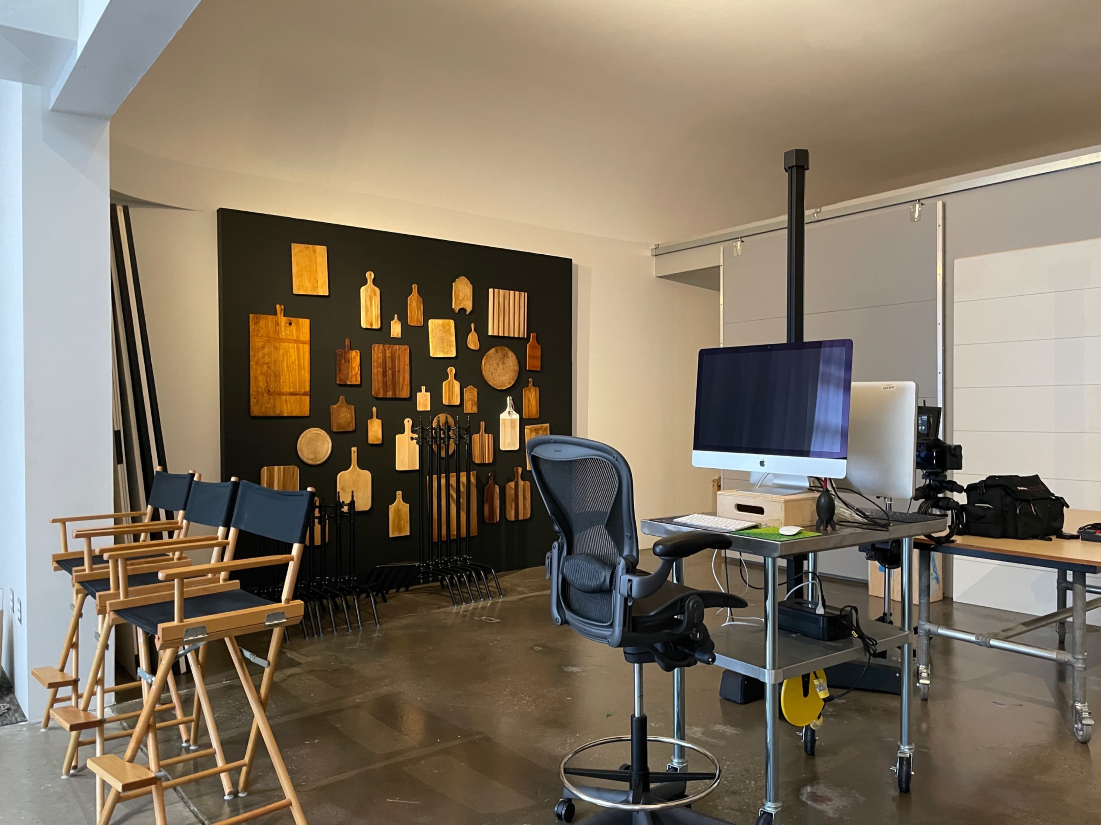 The image shows a modern office space with a large artwork on the wall made up of various wooden shapes, a desk with an iMac, and director's chairs arranged nearby.