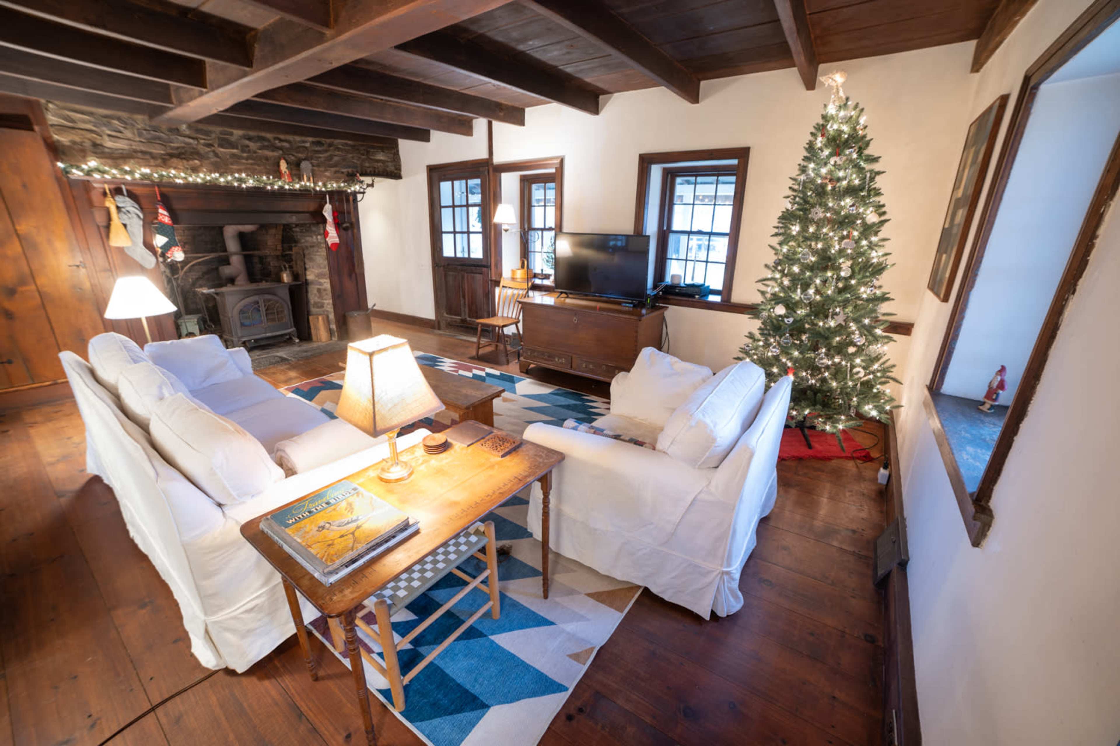 A cozy living room with white couches, a wooden table, a Christmas tree in one corner, and a stone fireplace.