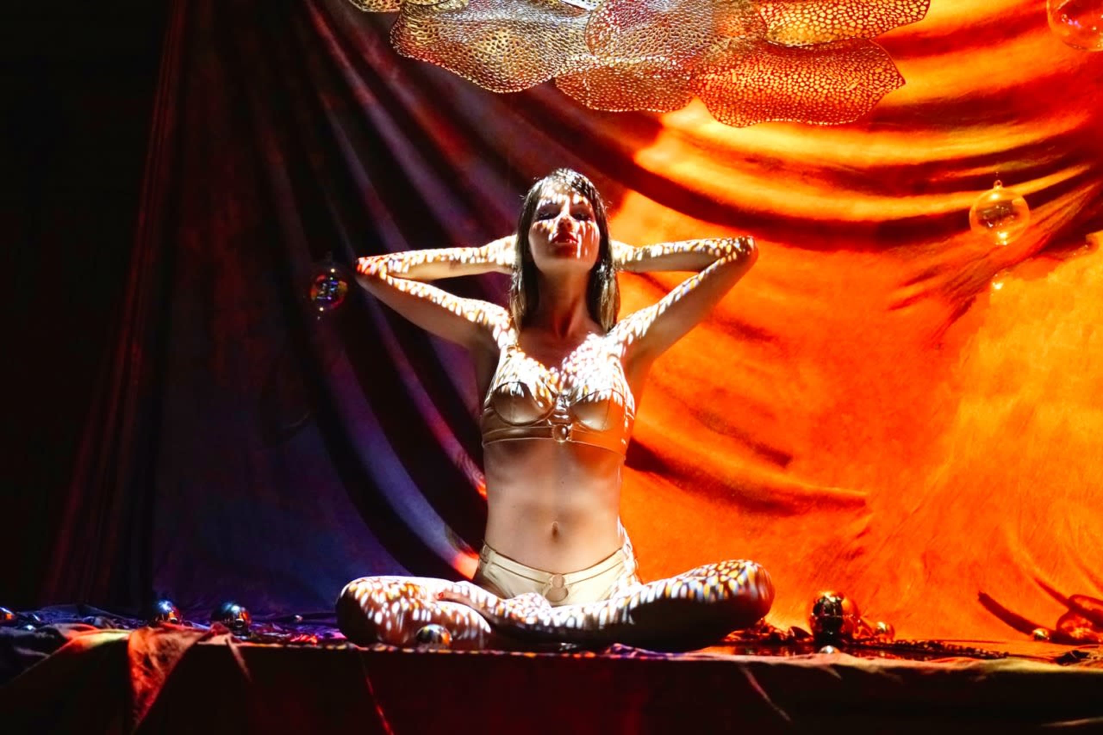 A woman in a metallic bikini sits cross-legged against a backdrop of colorful drapery, with light patterns projected onto her skin.