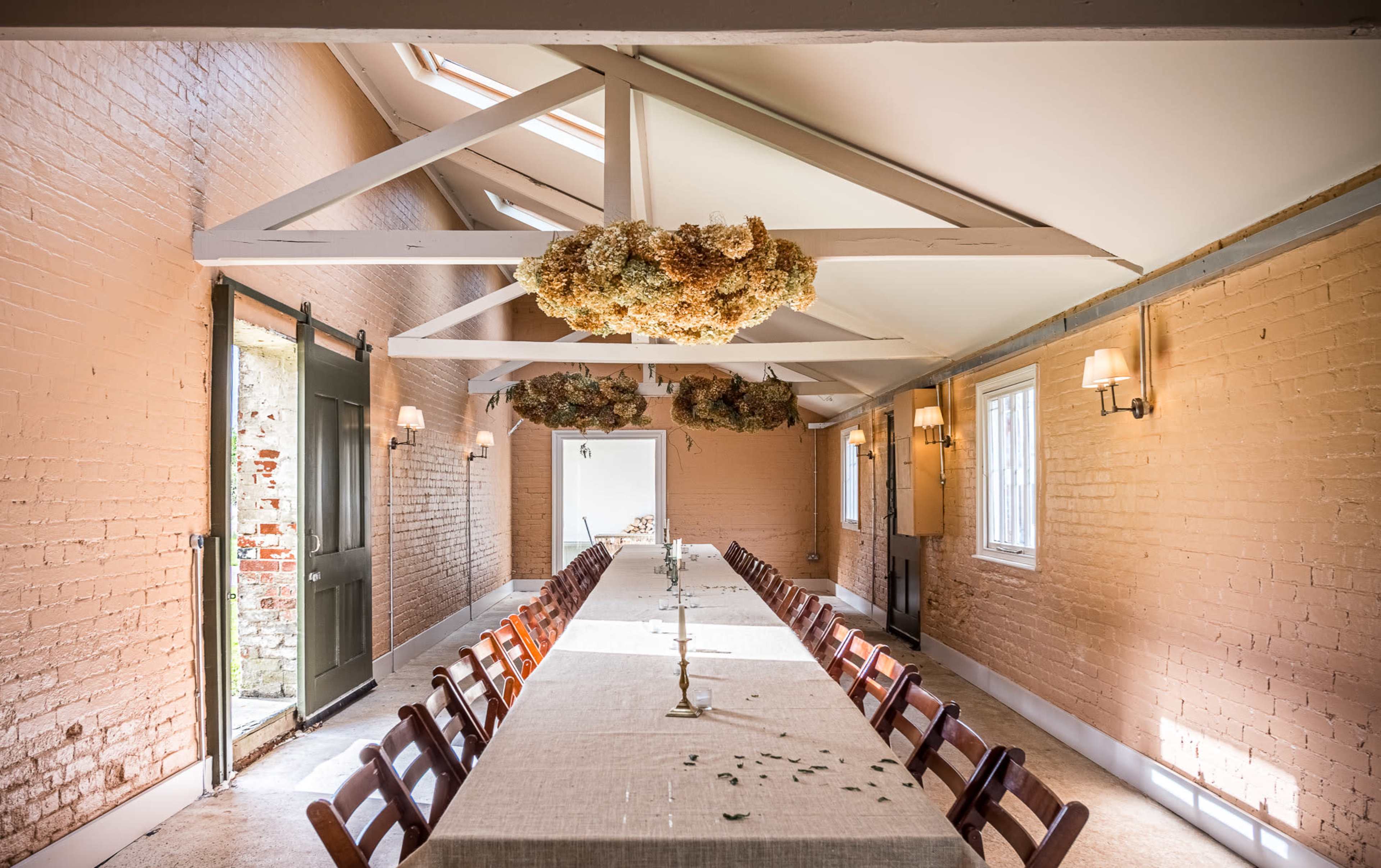 The image shows a long dining table set in a well-lit room with exposed brick walls and a wooden ceiling decorated with dried flowers.