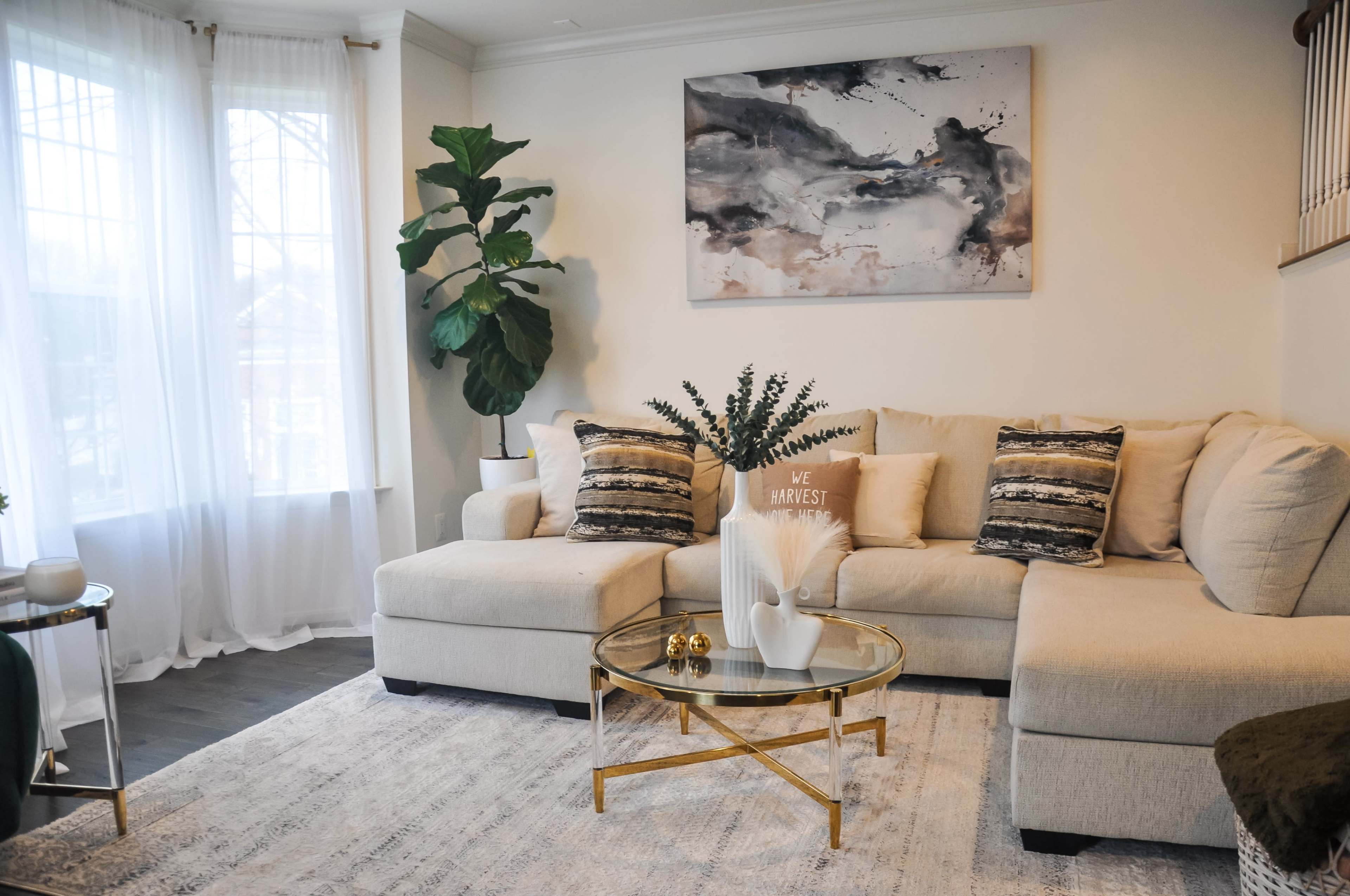 The image shows a modern living room with a beige sectional sofa, a glass coffee table, decorative pillows, and a large piece of wall art.