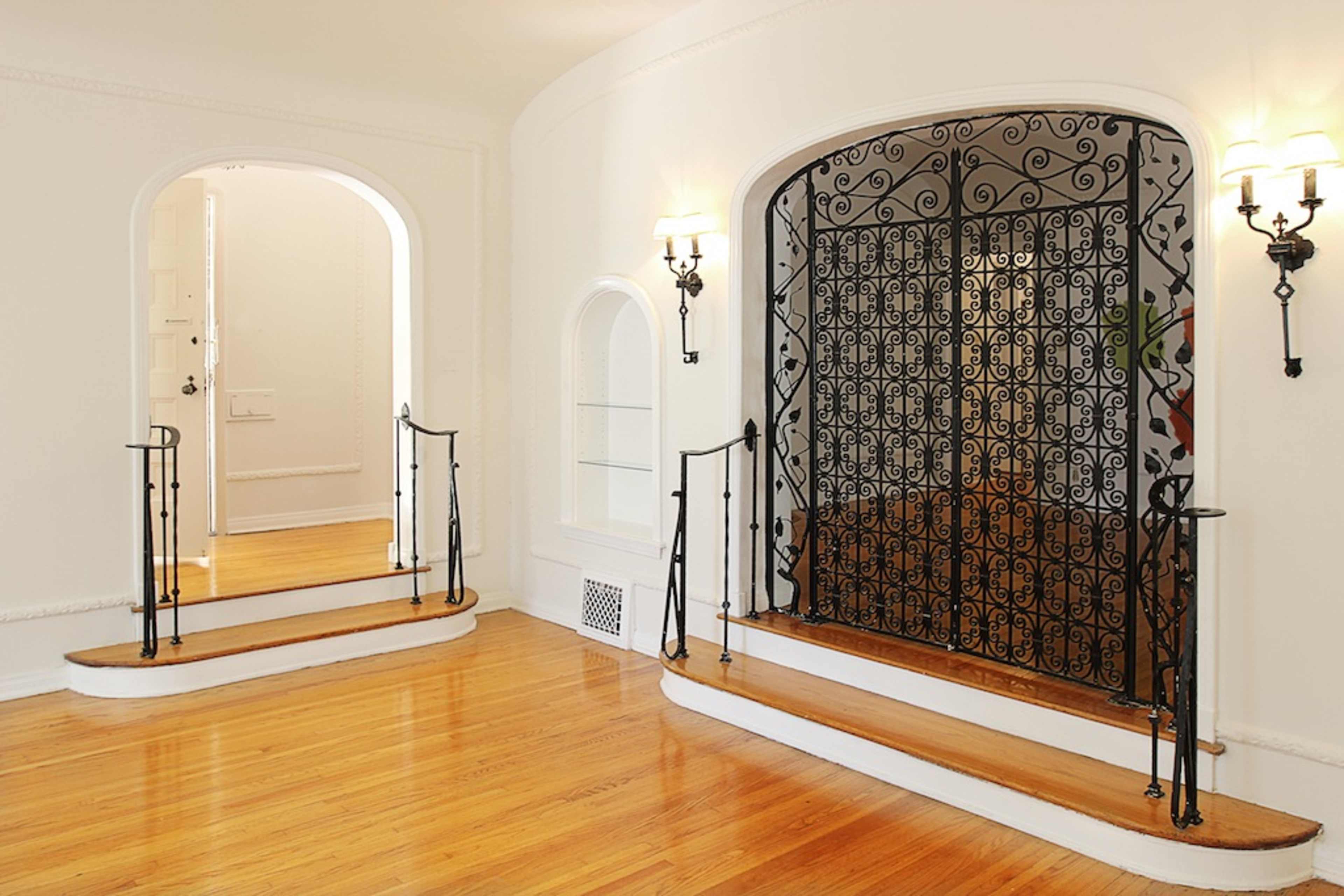 The image shows a spacious room featuring two wooden staircases leading to an ornate wrought iron gate, with warm wall sconces illuminating the space.