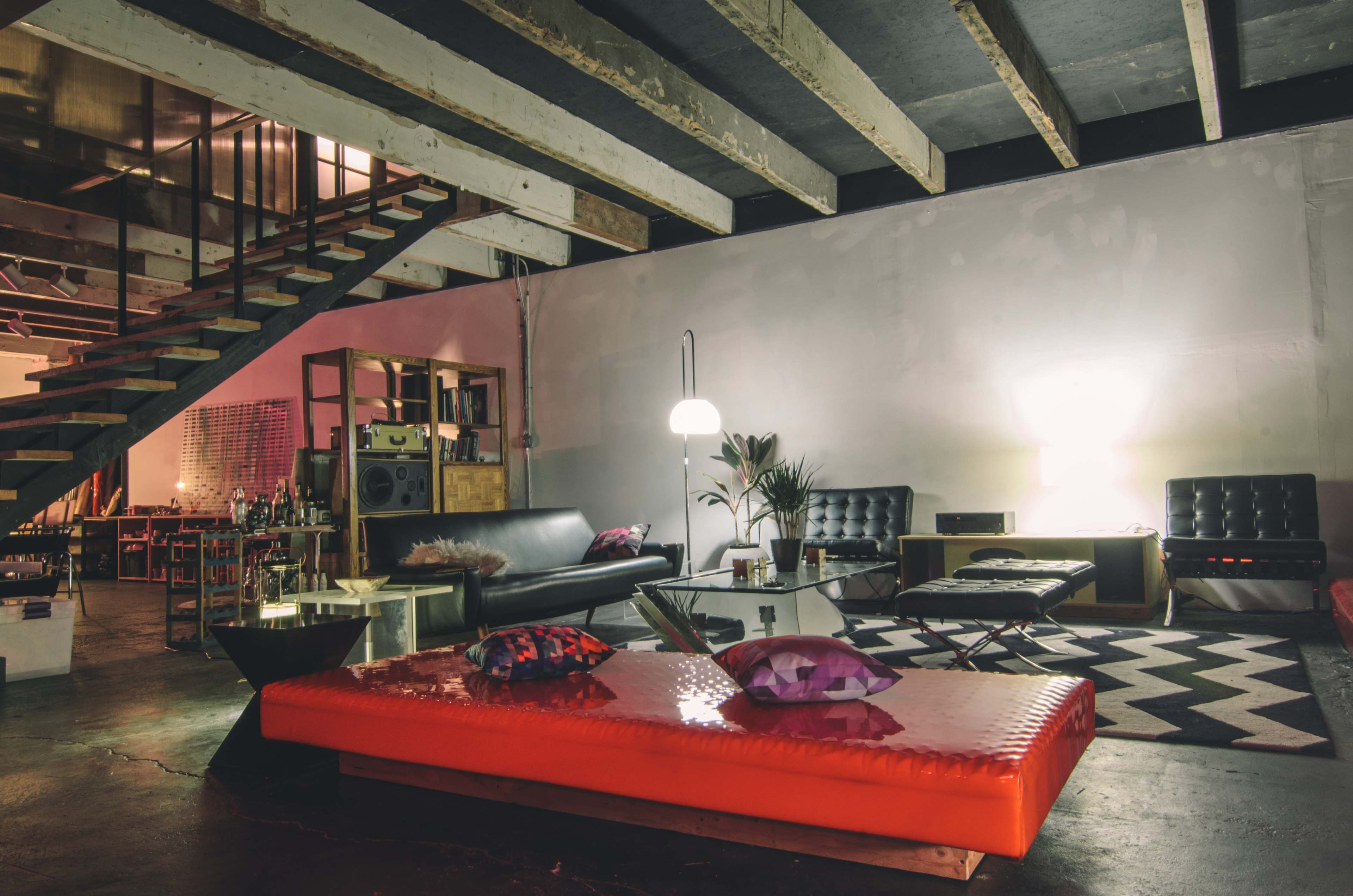 A modern, minimalist living space featuring an orange low table, black leather sofas, a patterned rug, and a staircase leading to an upper level.