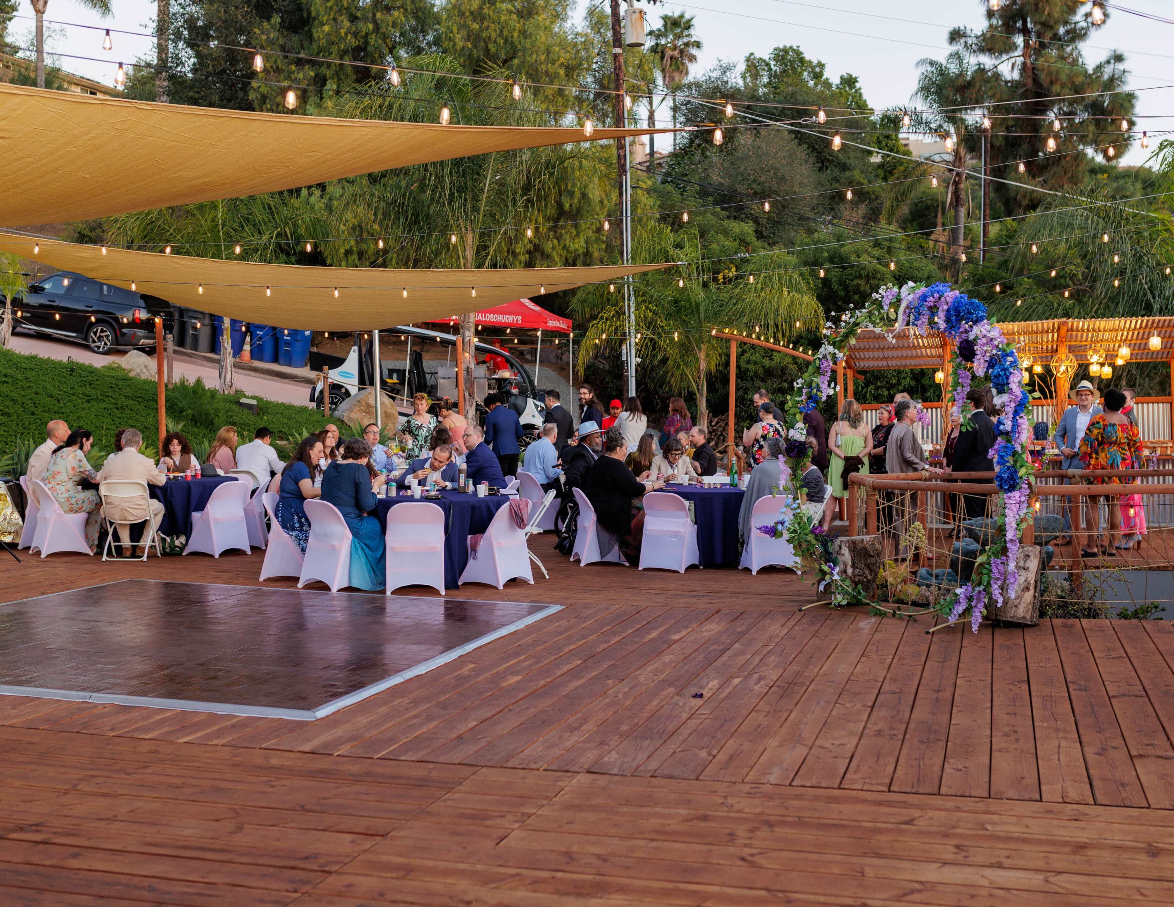 The scene depicts a lively outdoor event with guests seated at tables under large canopies, string lights illuminating the area, and a decorated entrance nearby.