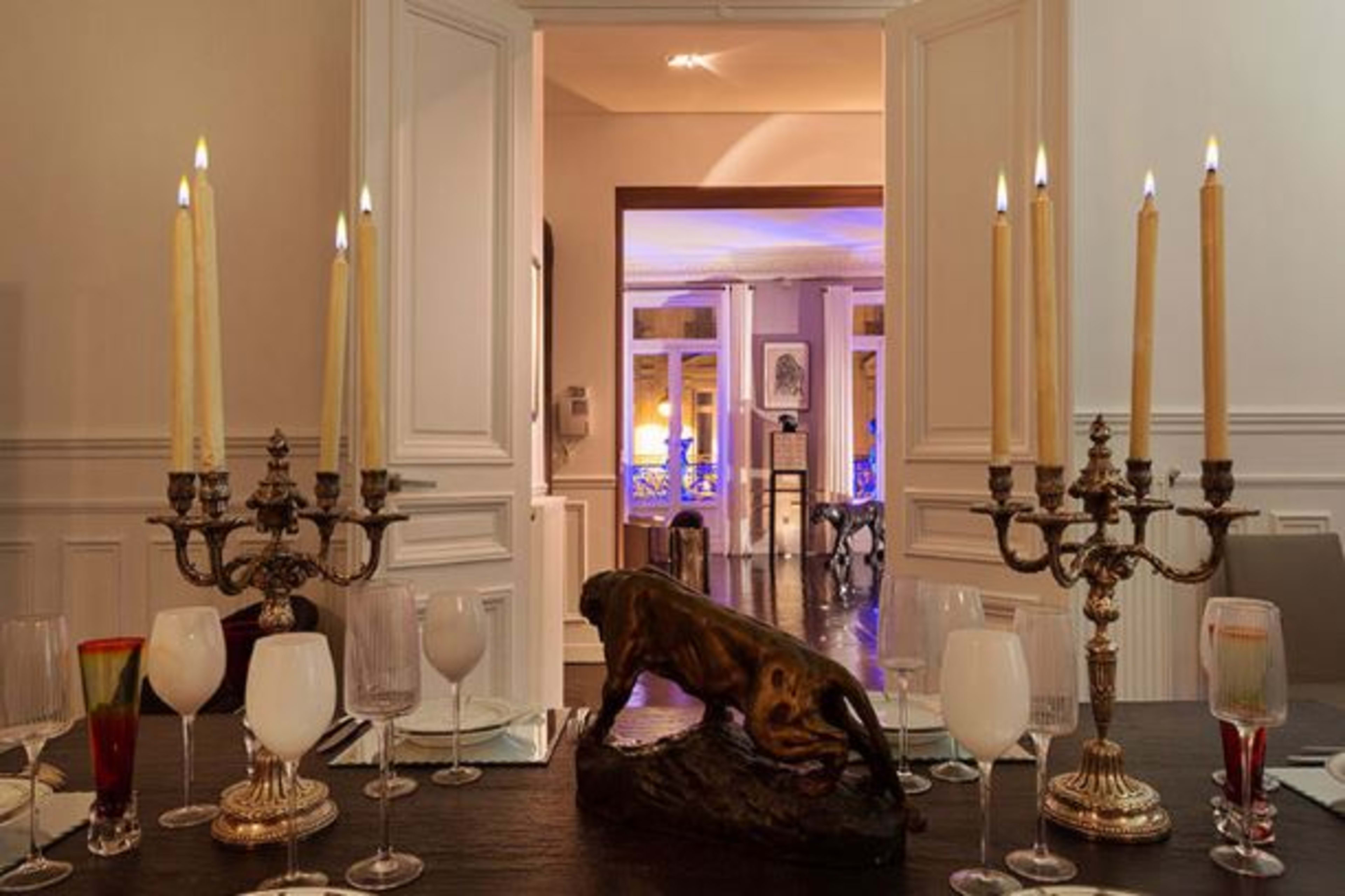 A dining table is set with candles and wine glasses, leading to a brightly lit room in the background.