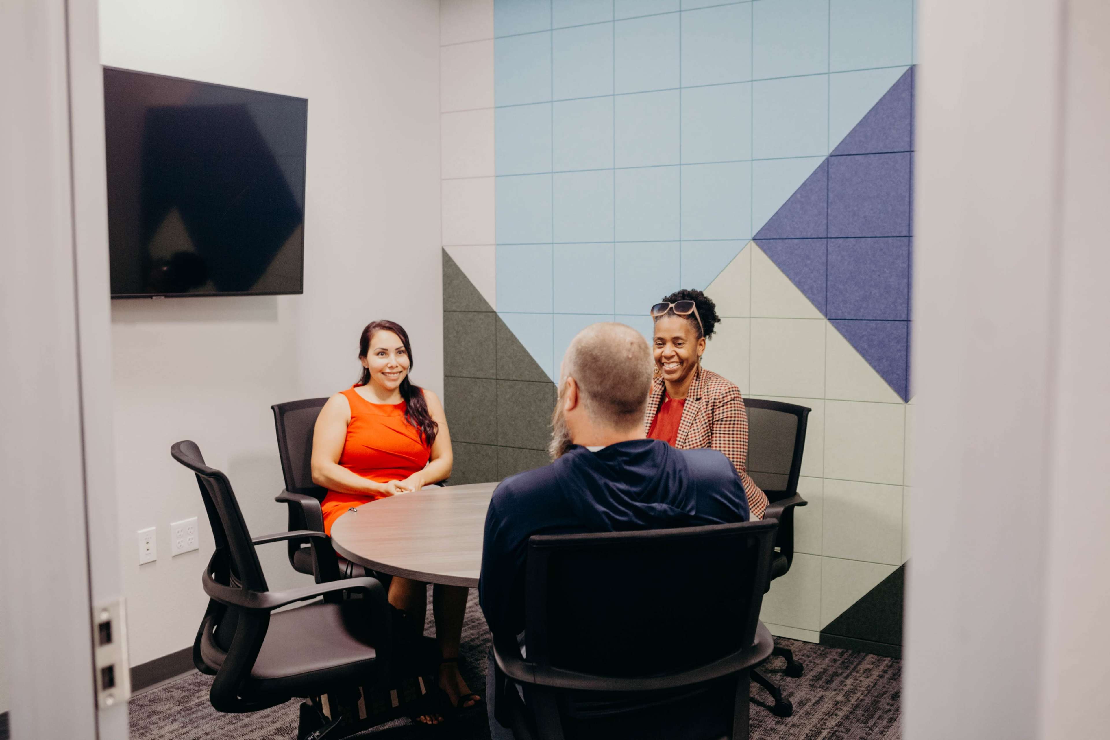 Three people are seated around a circular table in a small meeting room with a colorful wall.