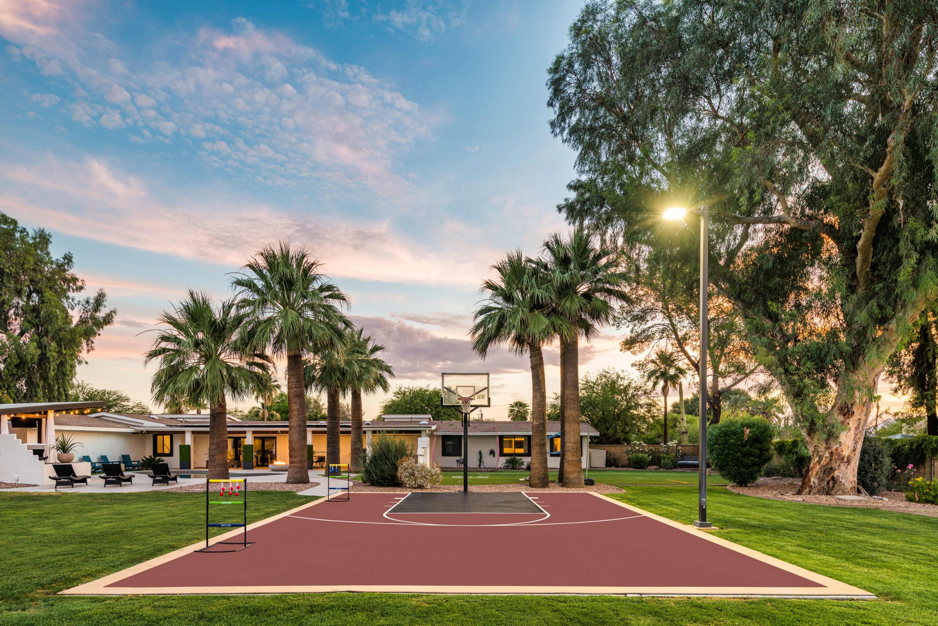 A basketball court surrounded by palm trees and a well-maintained lawn is set in front of a modern house under a colorful sky.