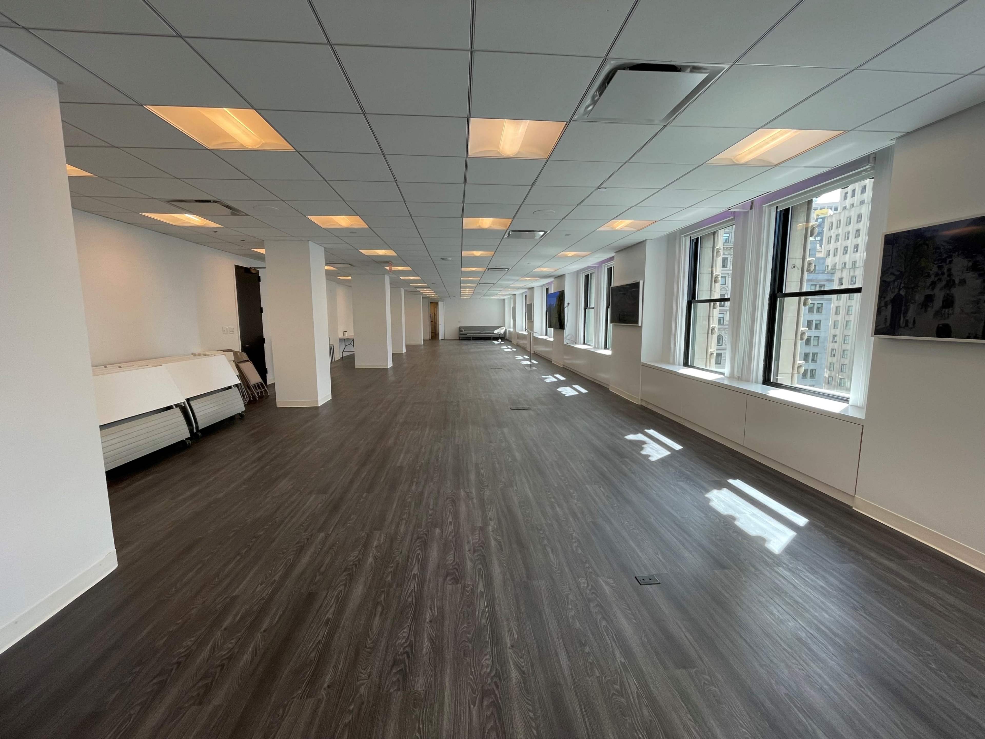 A spacious, unoccupied office interior with gray flooring, large windows, and several mounted screens along the walls.