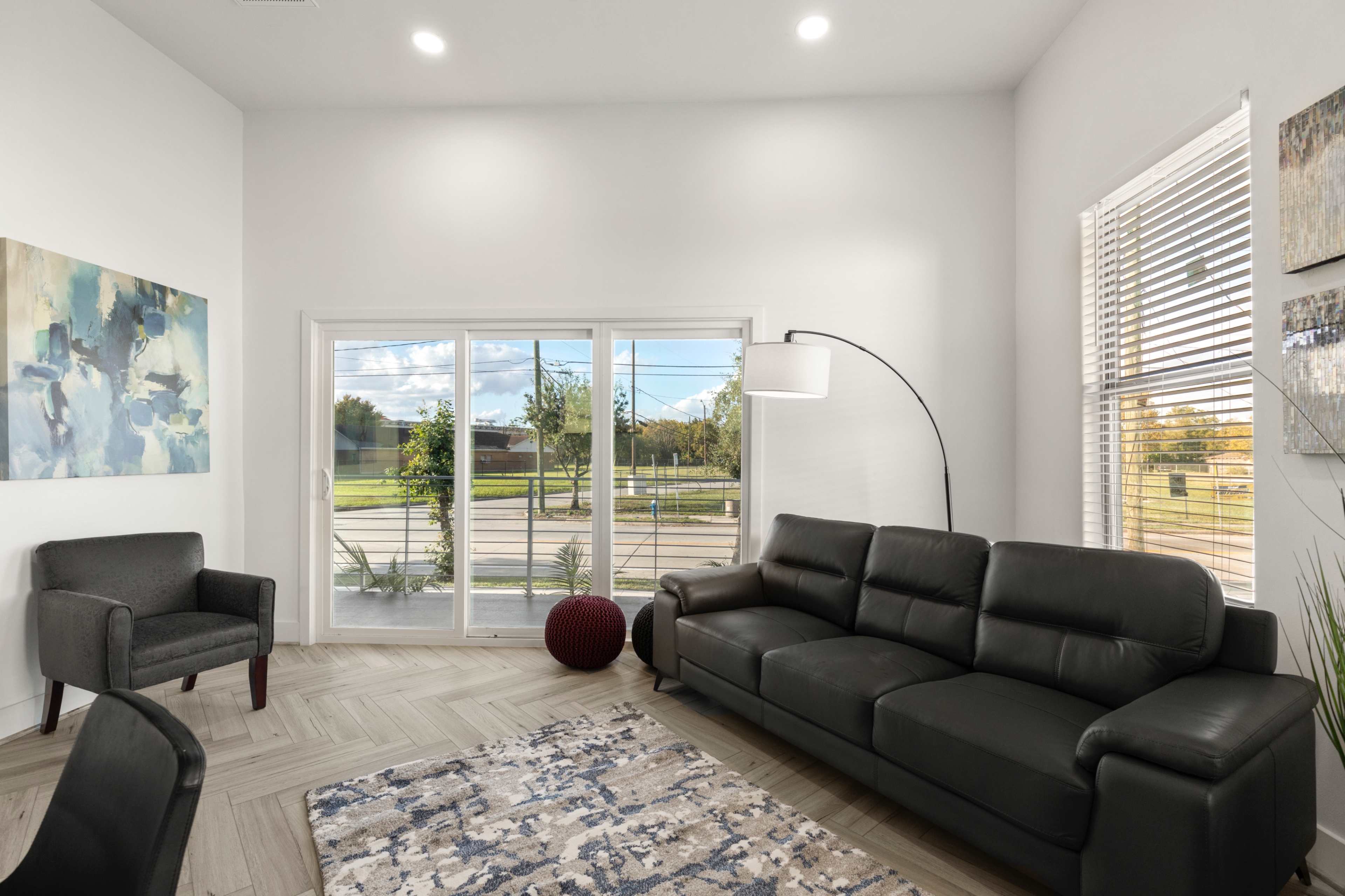 A modern living room features a dark leather sofa, an armchair, a floor lamp, and large windows with outdoor views.