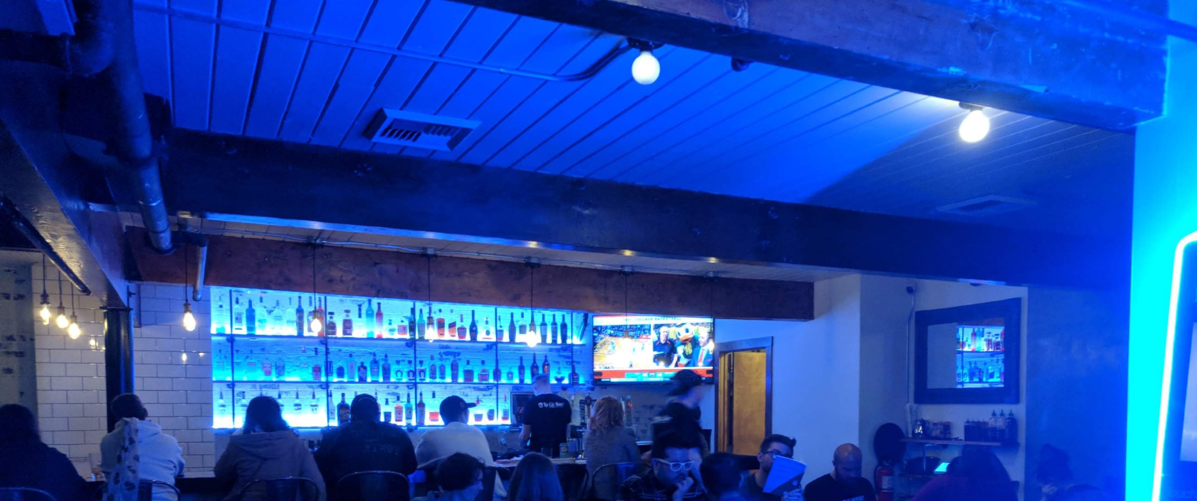 A bar interior with patrons seated in front of a backlit shelf of liquor bottles, illuminated by blue lights and featuring multiple screens displaying TV broadcasts.