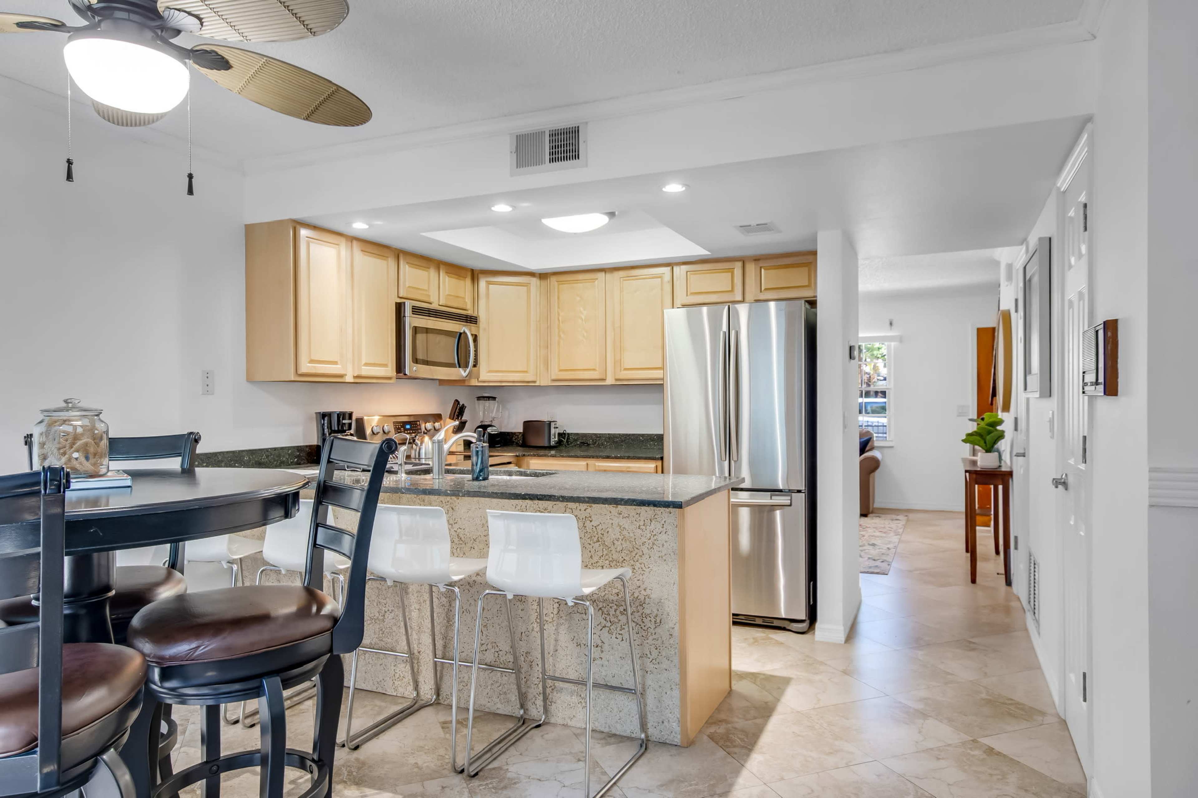 A modern kitchen with light wood cabinetry, a granite countertop, and stainless steel appliances, featuring a breakfast bar with white stools.