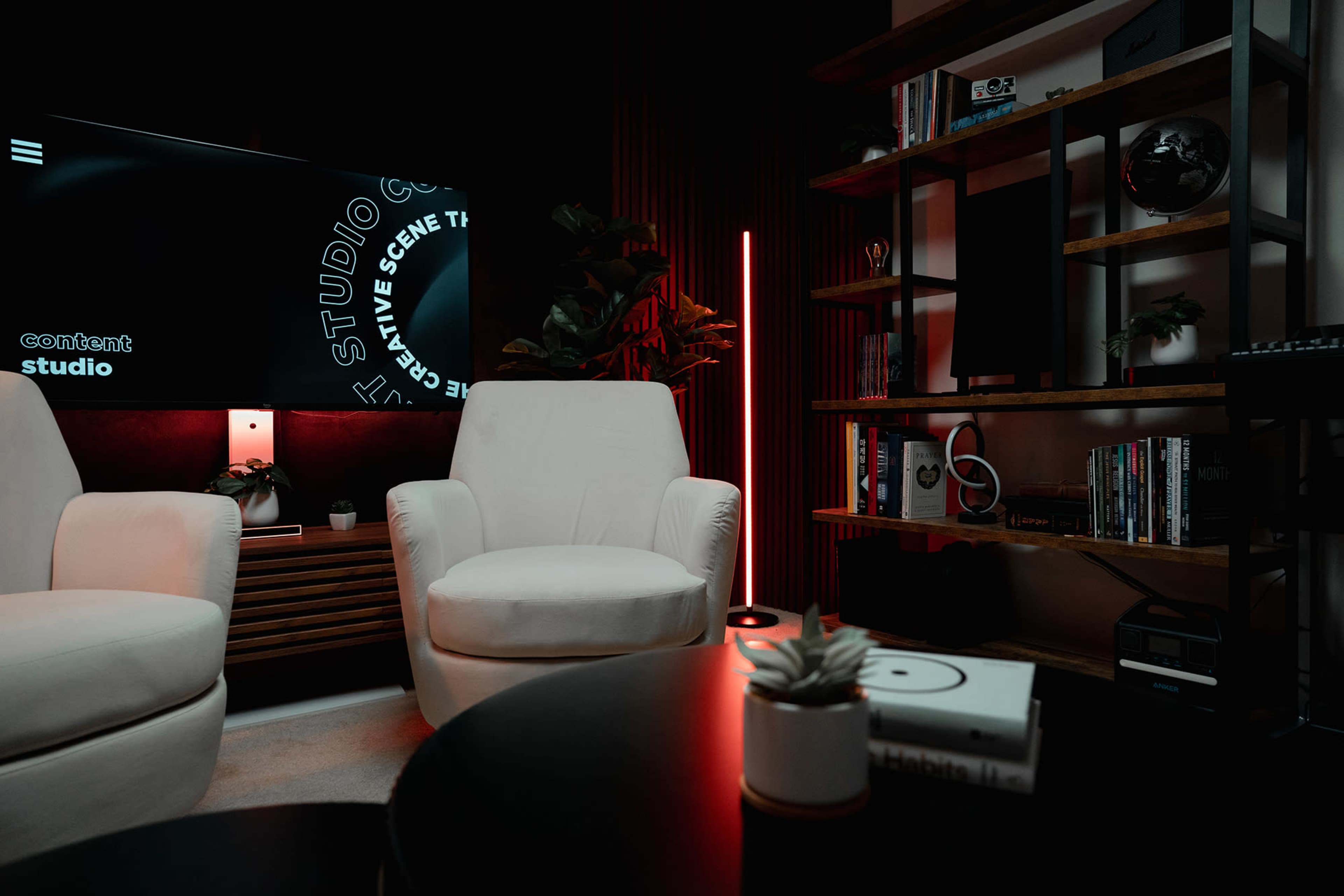 A modern content studio features two white armchairs, a large television on the wall, and a bookshelf filled with media items.