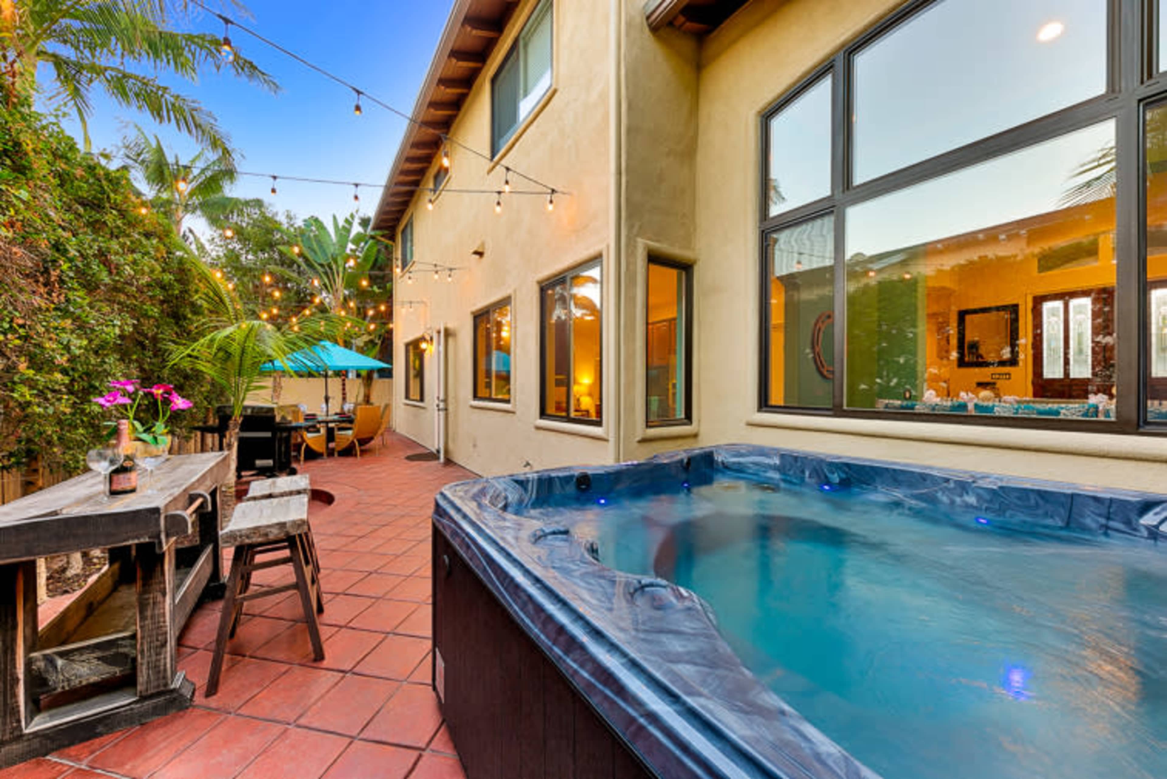 A hot tub is positioned next to a patio area with string lights and outdoor furniture adjacent to a house surrounded by palm trees.