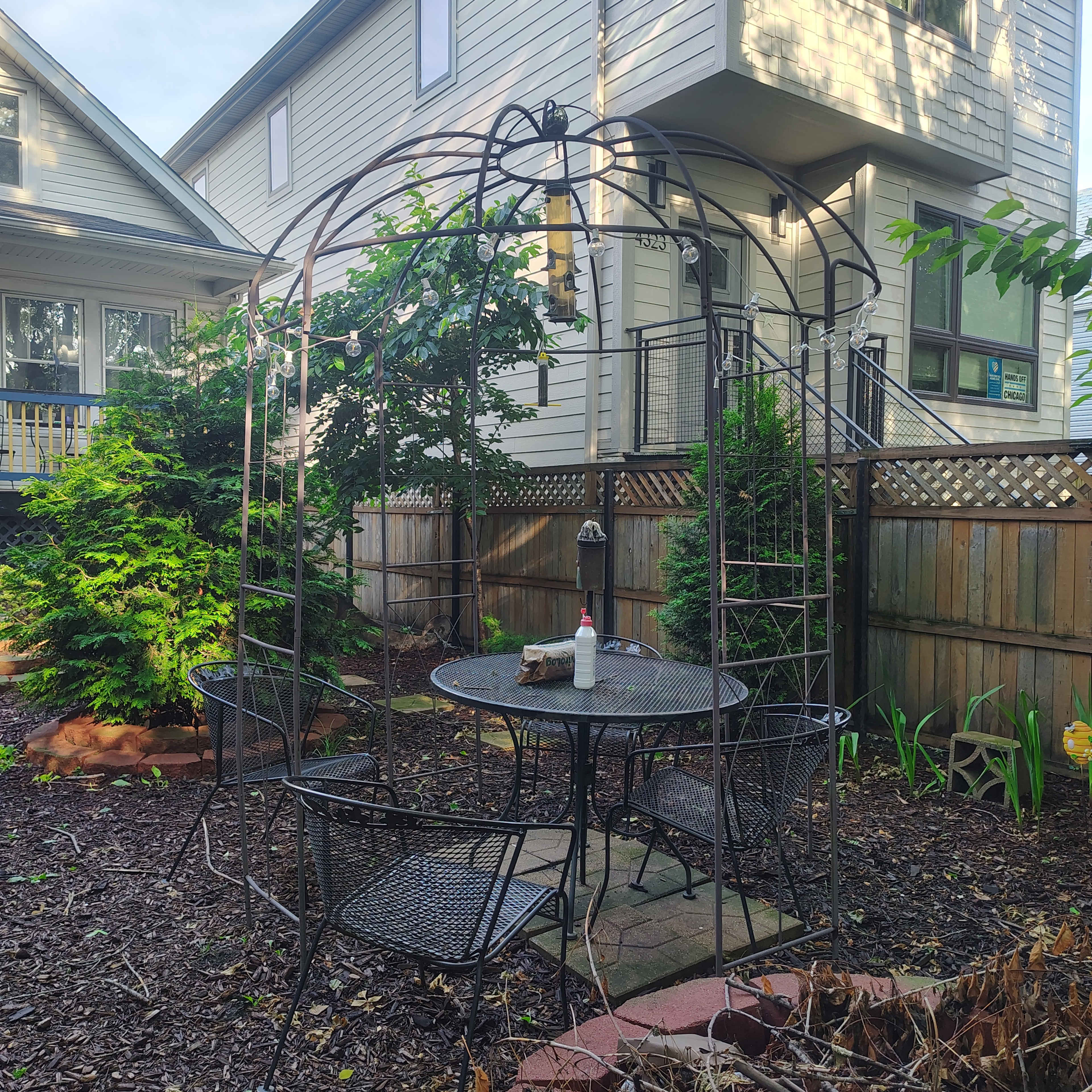 A metal gazebo with a table and chairs is situated in a garden surrounded by fences and greenery.