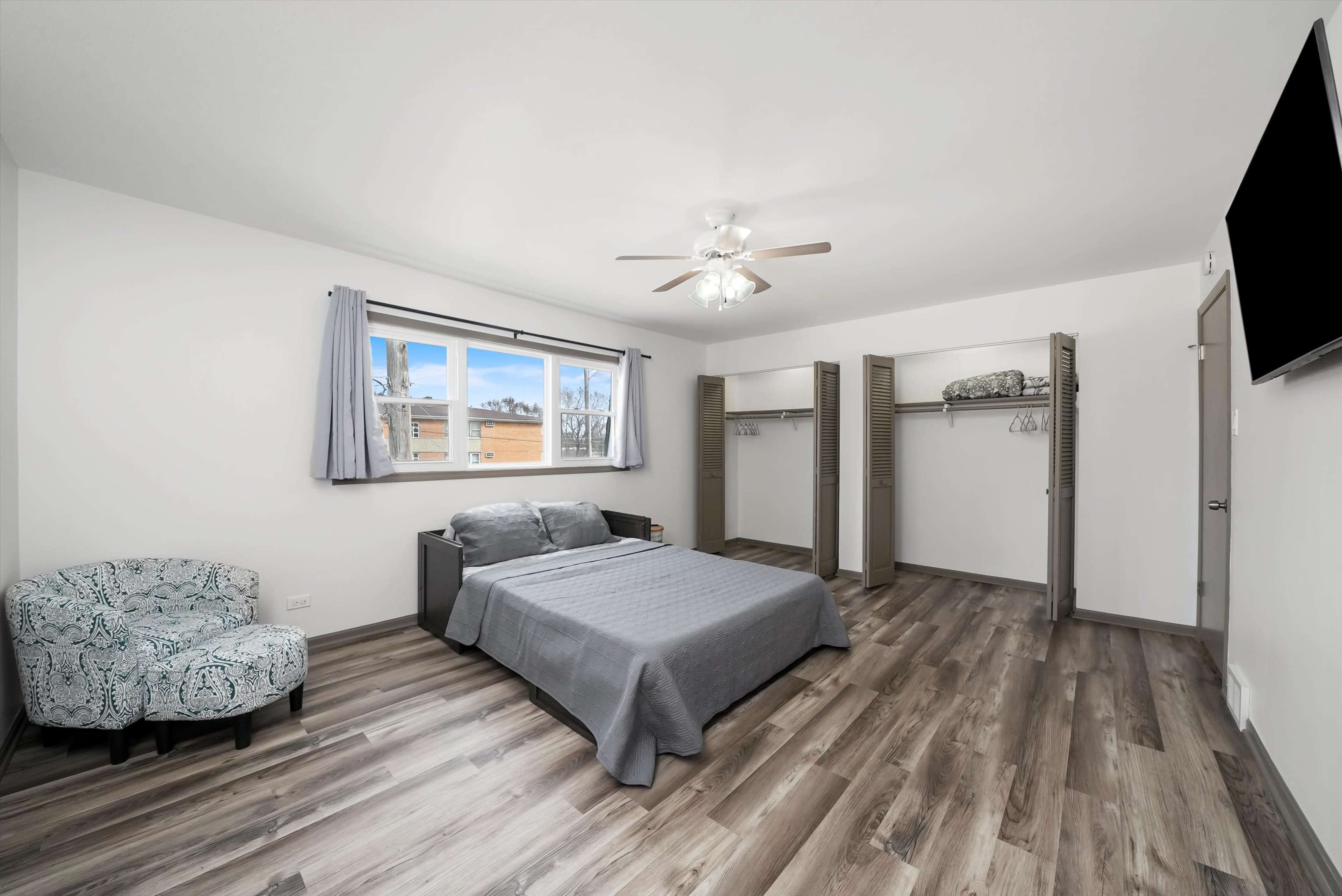 A minimalist bedroom with a bed, a ceiling fan, a window, and two open closet spaces.