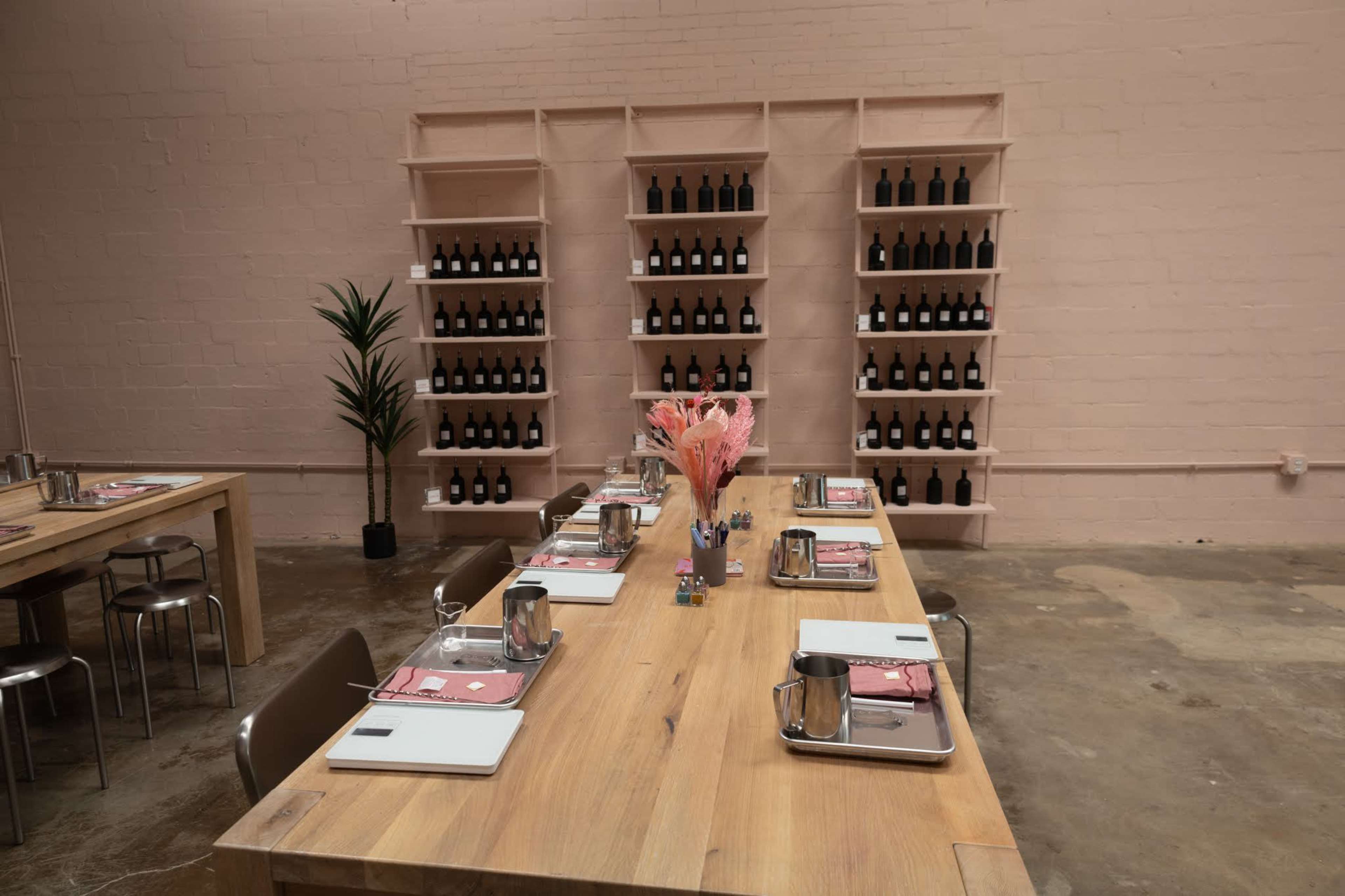 A spacious dining area with a large wooden table set for multiple guests, adorned with a bouquet of pink flowers, and a wall shelf lined with bottles in the background.