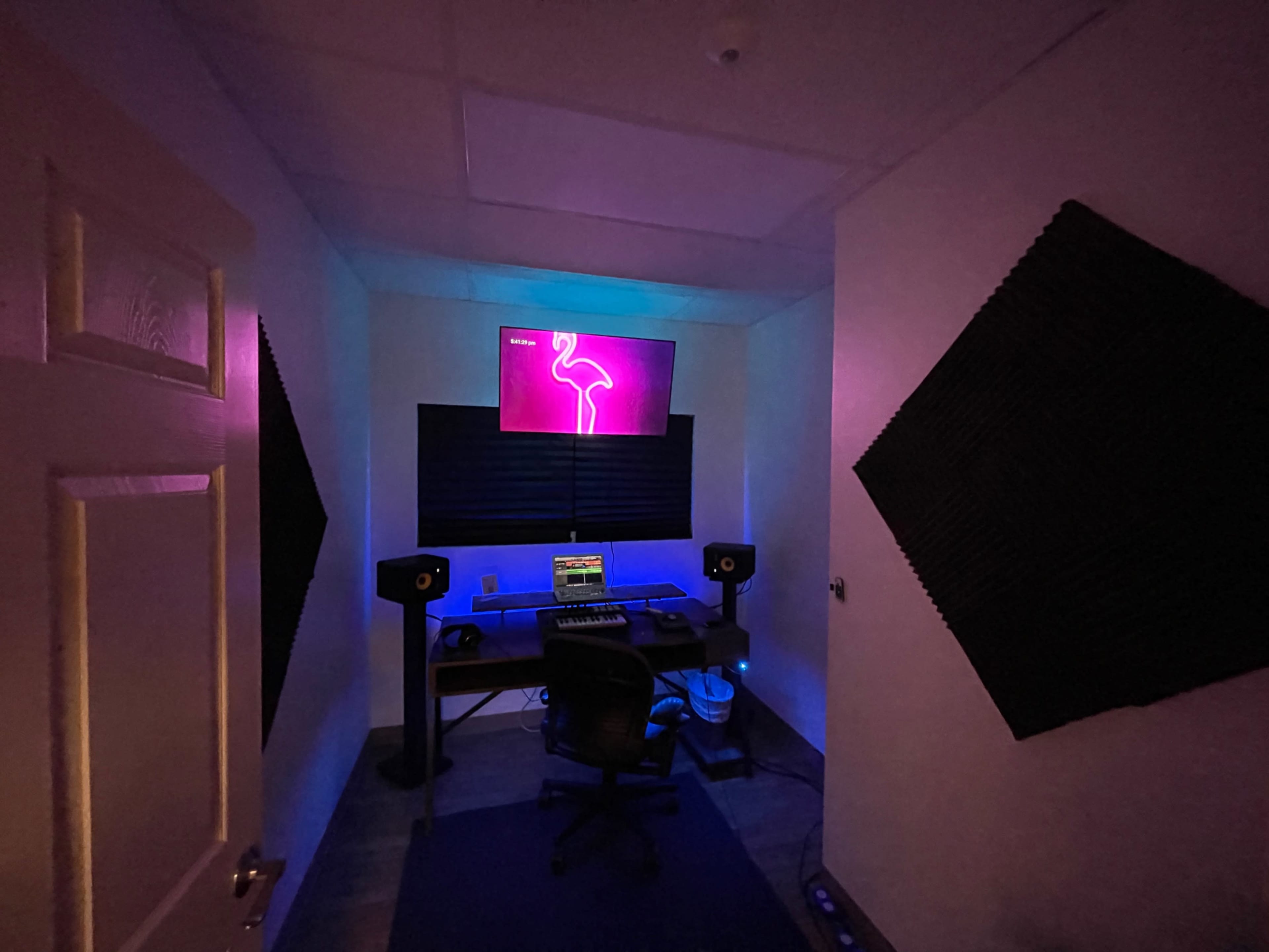 A music production room with a desk, computer, and speakers, illuminated by purple and blue lighting.