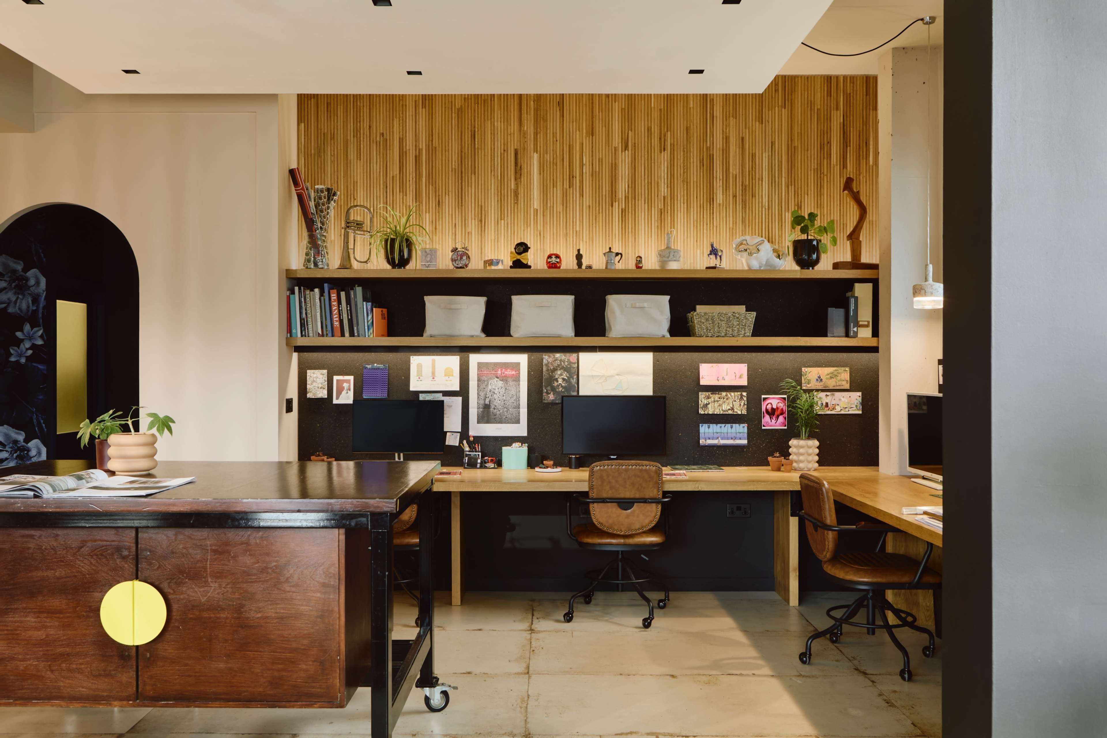 A modern office space with two desks equipped with computers and a wooden wall featuring shelves displaying various decorative items and plants.