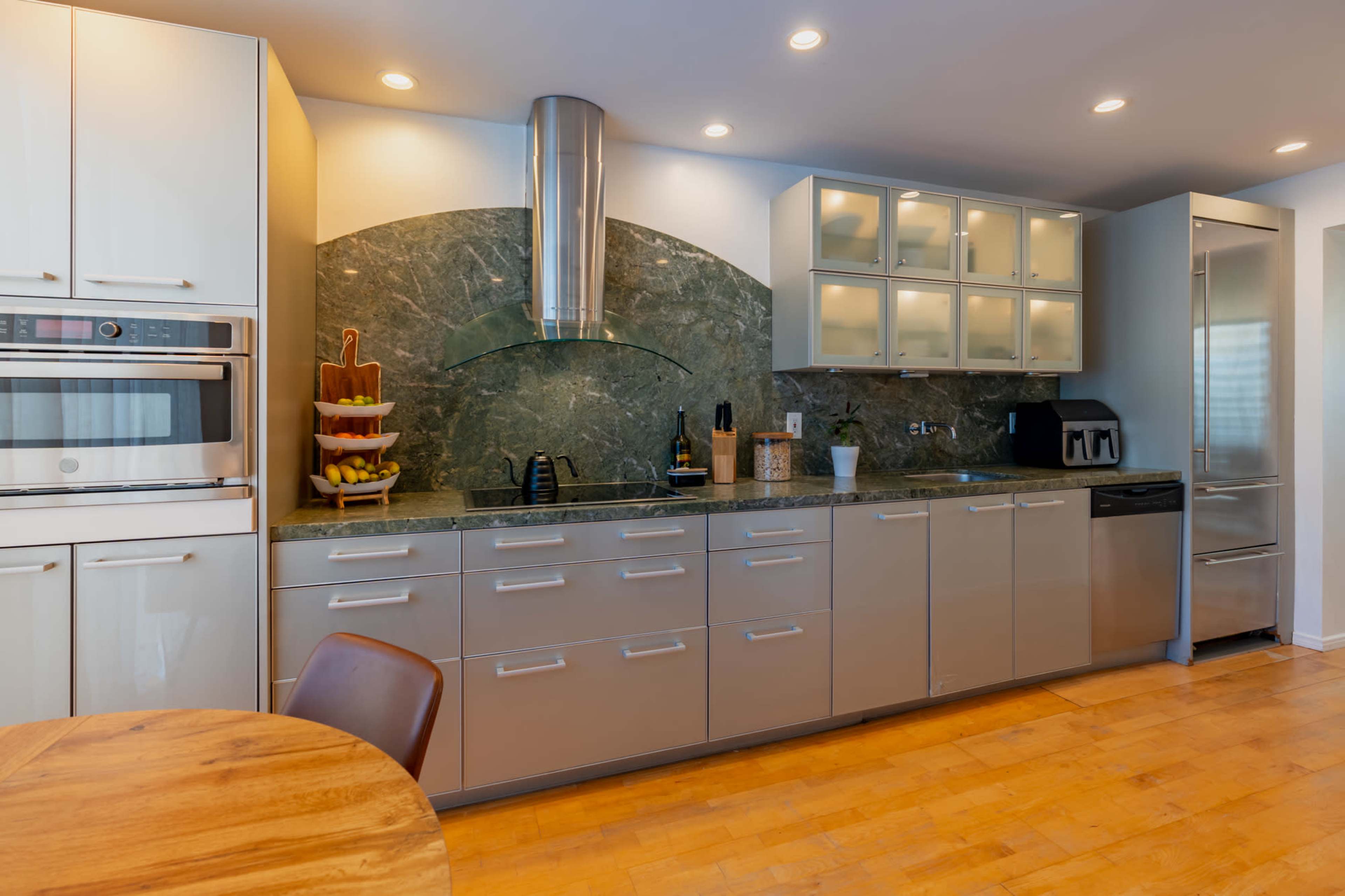 The image shows a modern kitchen with sleek stainless steel cabinets, a green marble backsplash, and a circular hood above the stove.