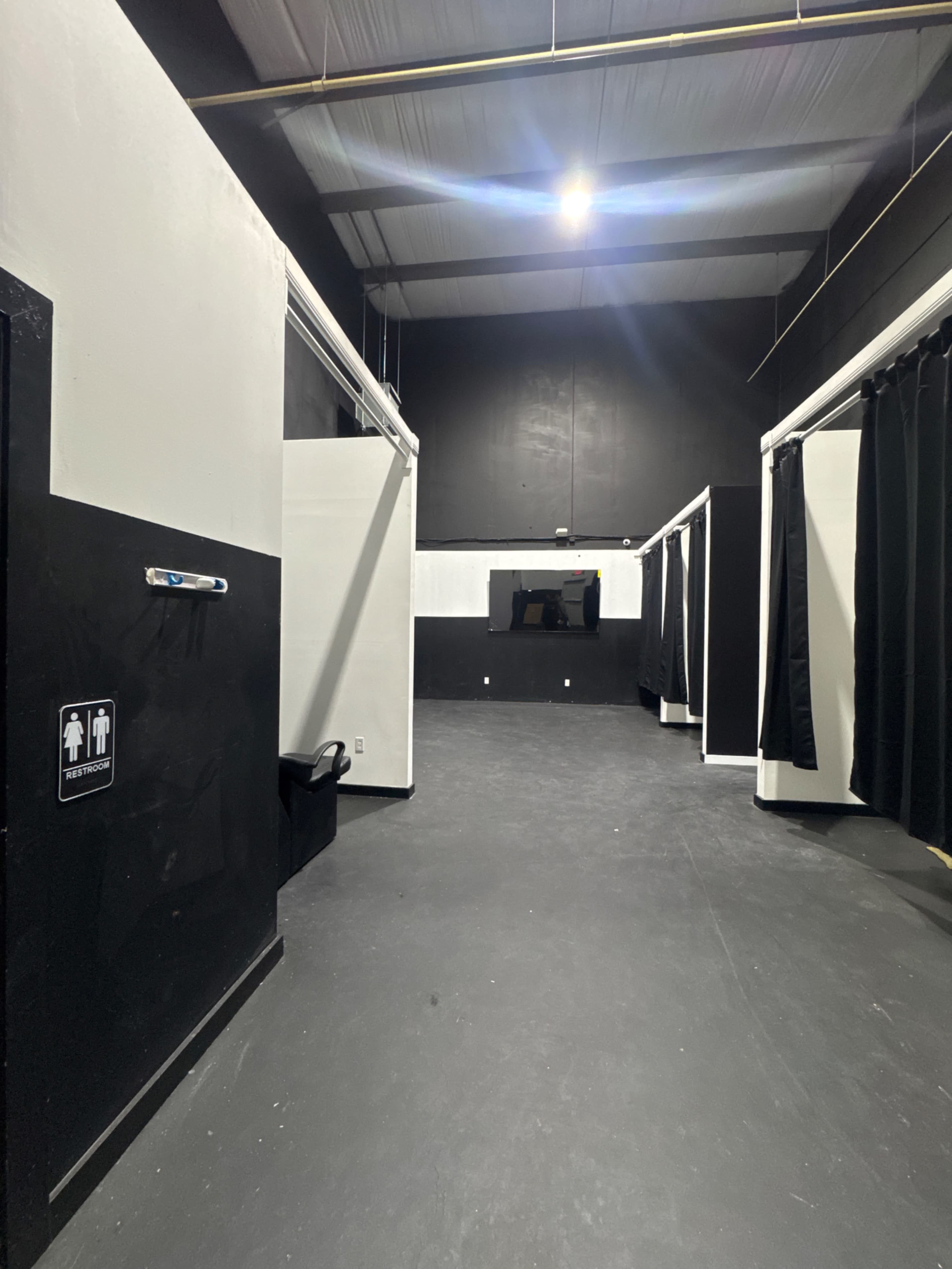 The image shows a spacious, dimly lit interior with black and white walls, featuring several black curtains and a door on the left marked with a restroom symbol.